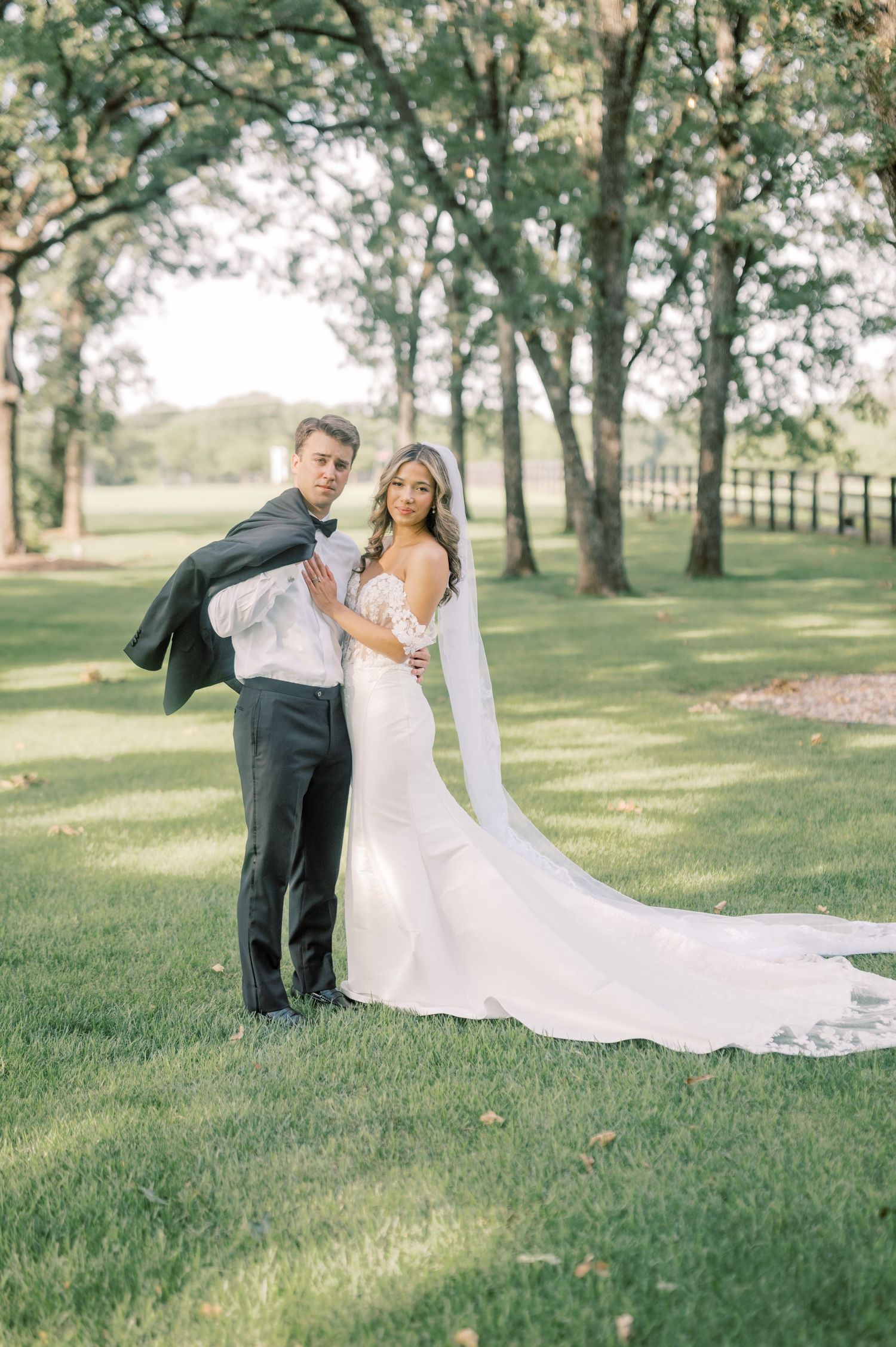 Bride in fitted lace gown with long veil and groom in tuxedo posing together on lush green lawn.