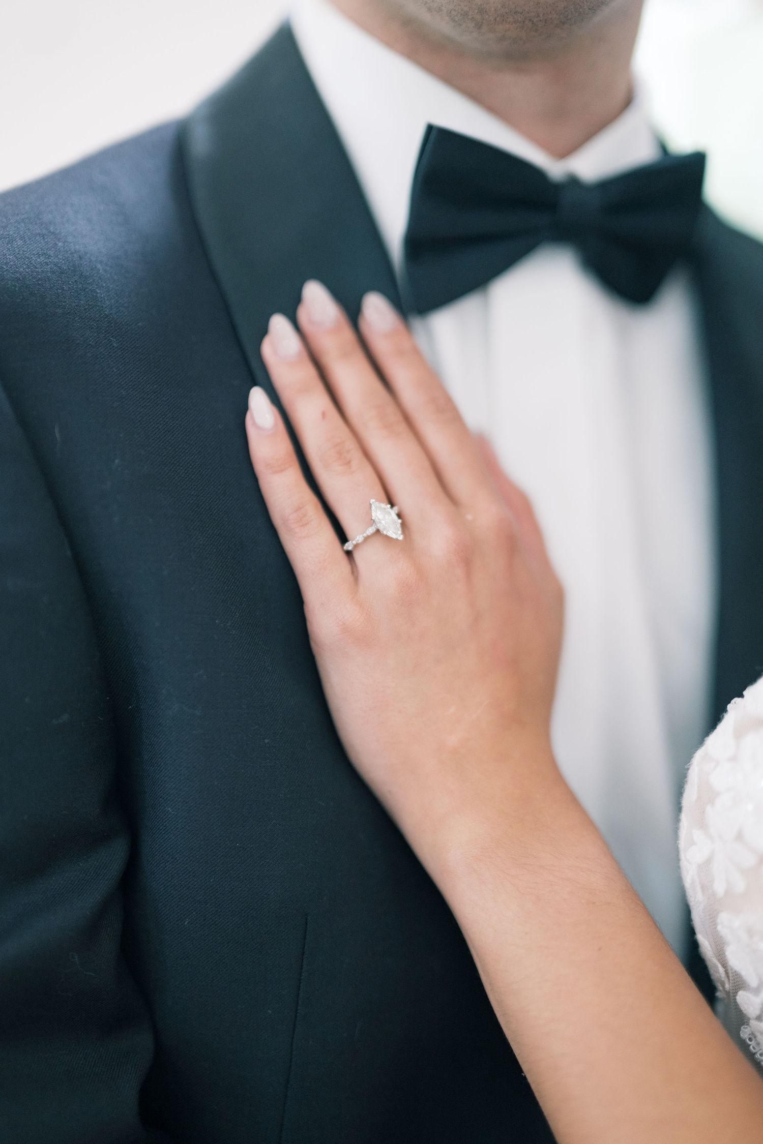 Close-up of bride's hand displaying a marquise diamond engagement ring resting on groom's dark tuxedo.