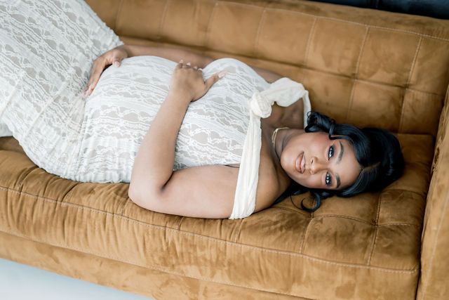 Pregnant woman in white lace dress lying on brown velvet sofa, cradling baby bump during maternity photo session.