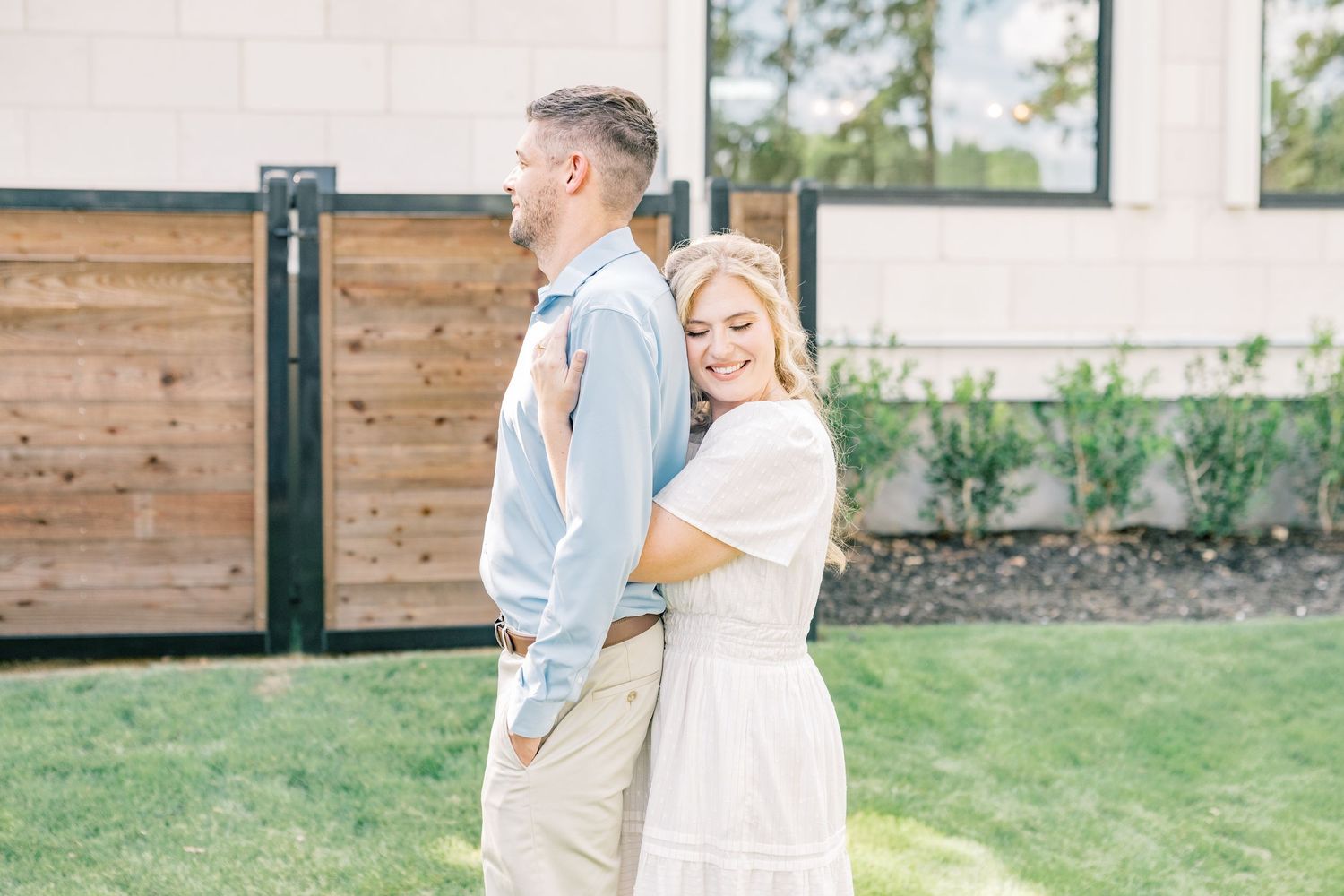 A smiling woman in a white dress embraces a man in a light blue shirt outdoors near a modern building.