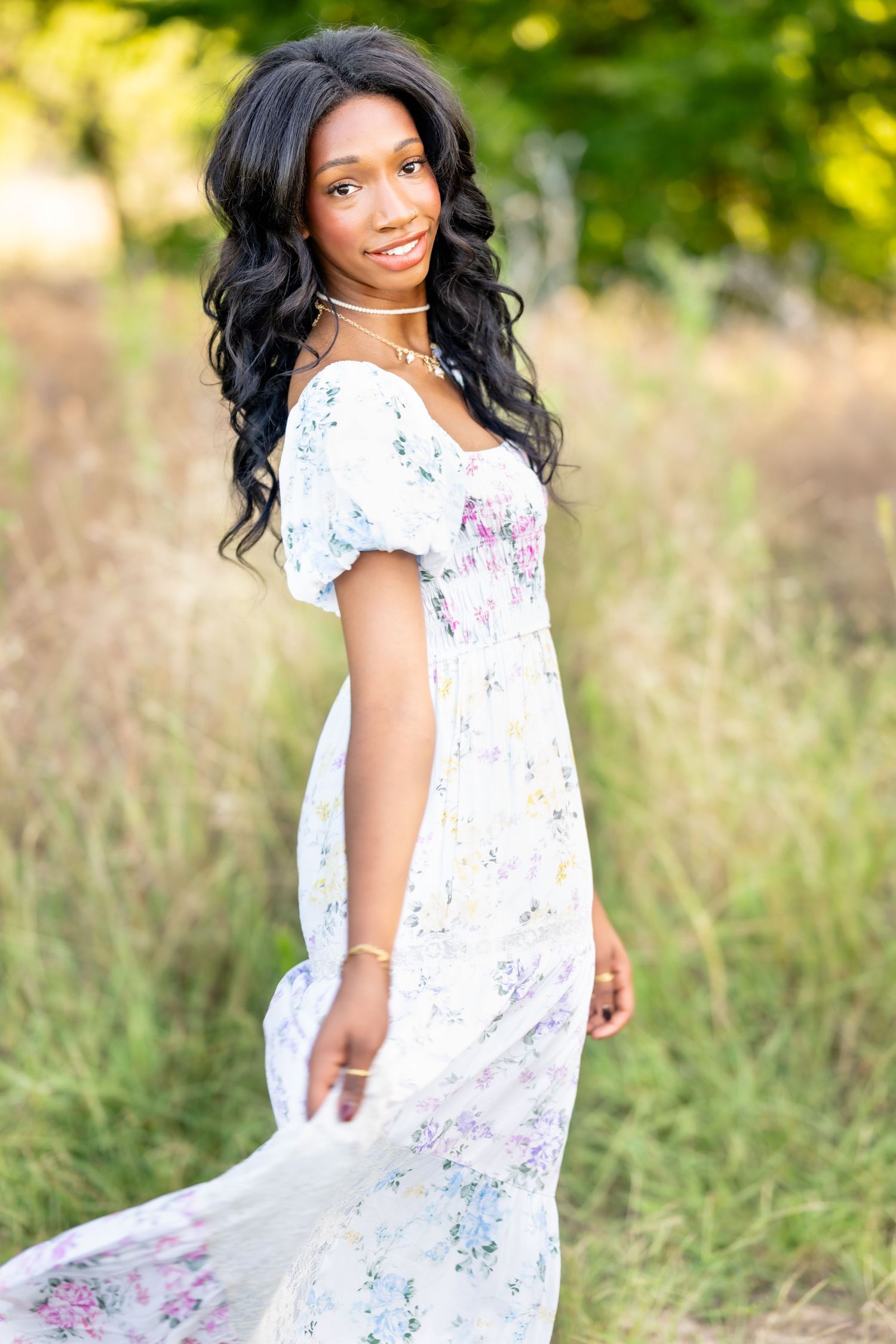 Young Black woman in white floral maxi dress poses outdoors in a field with lush green trees in background.