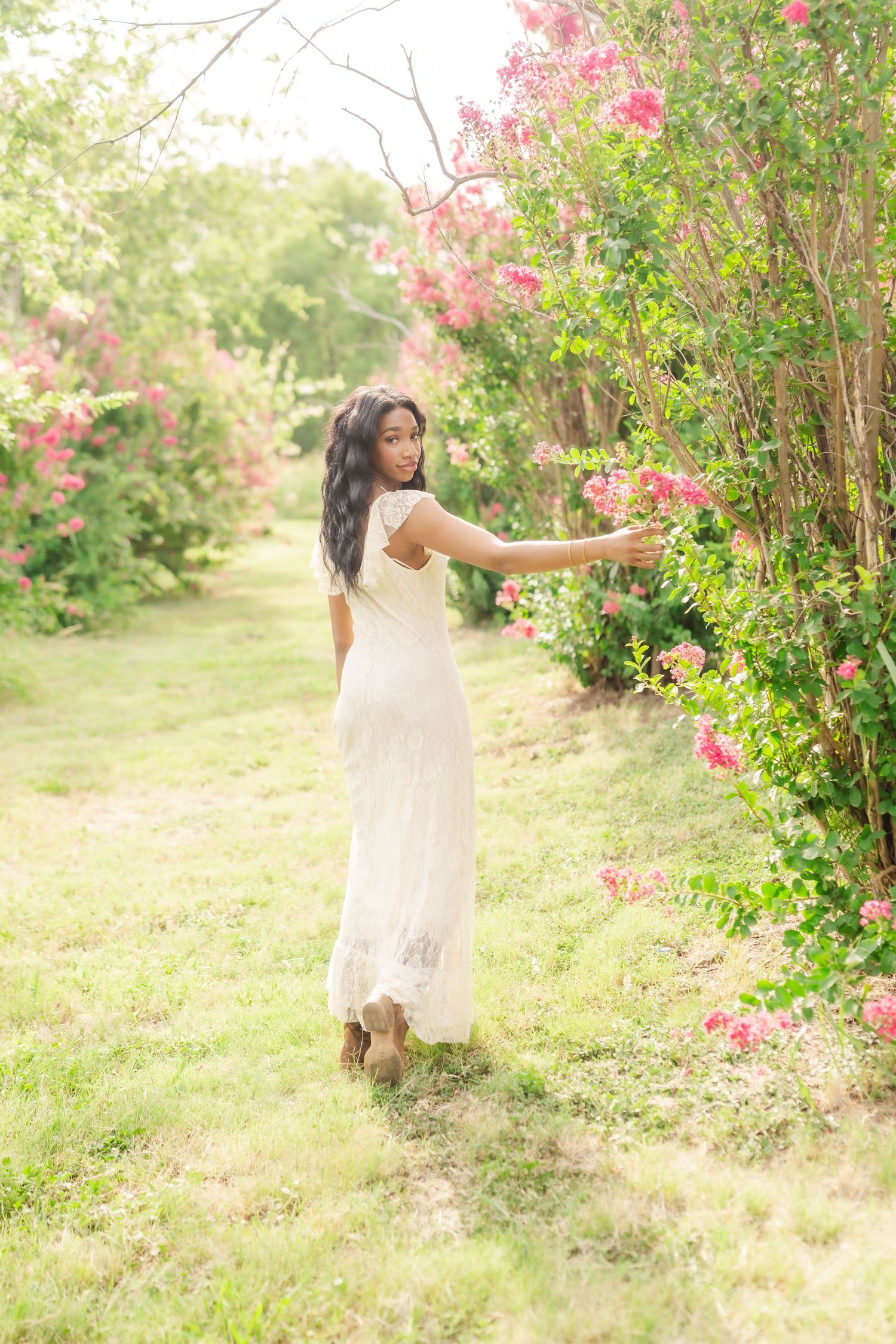 Woman in white dress reaching toward pink roses in a lush garden pathway.