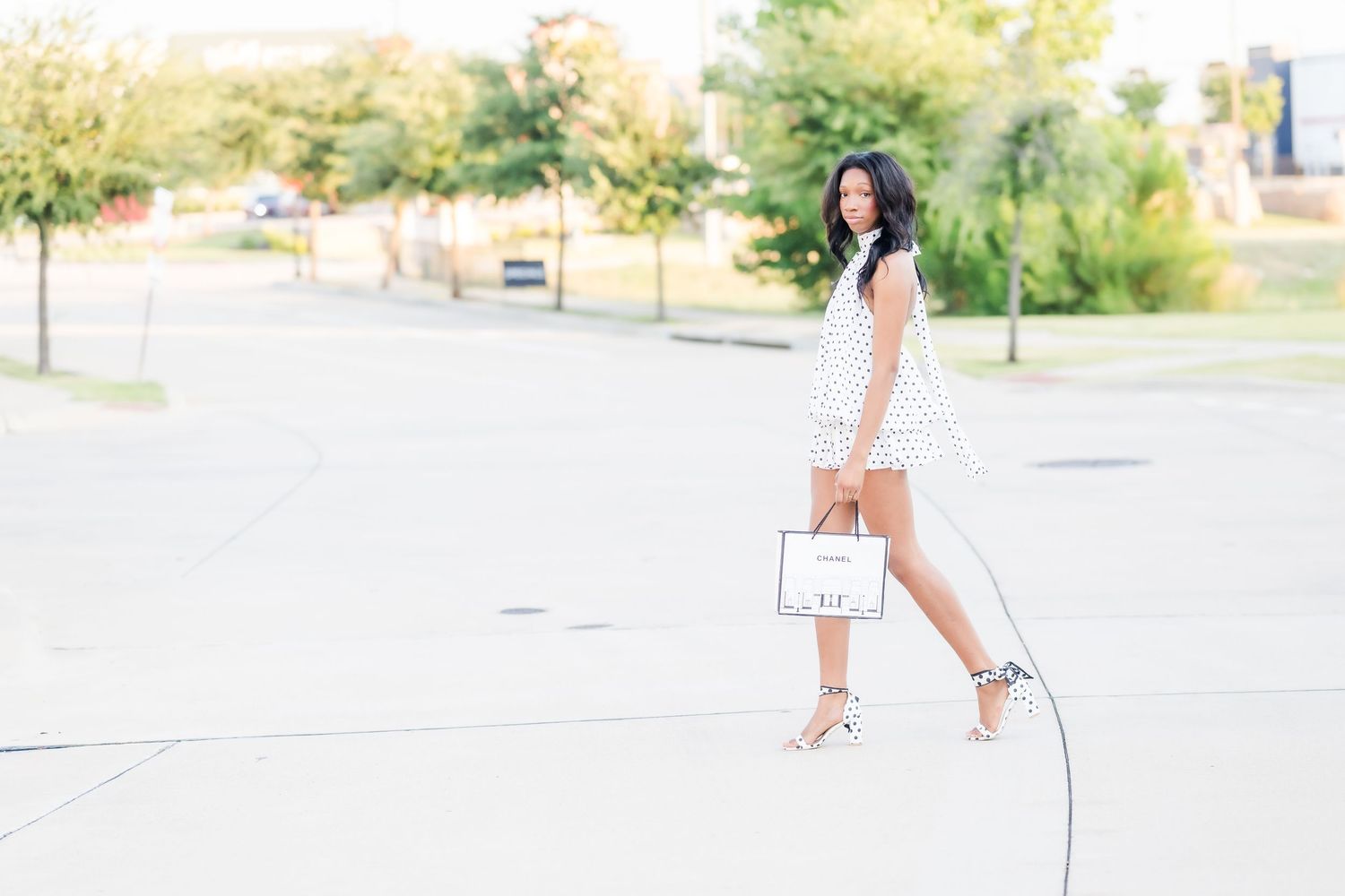 Woman in white polka dot dress holding a white handbag, posing outdoors on a sunny day.