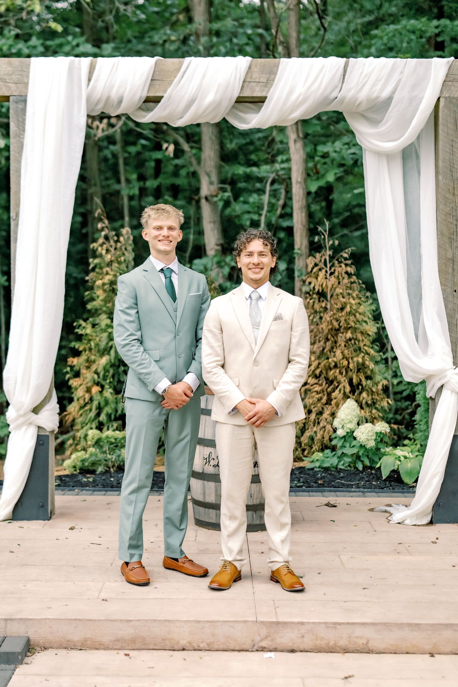 Two grooms in suits stand smiling under a white draped outdoor wedding arch surrounded by lush greenery.