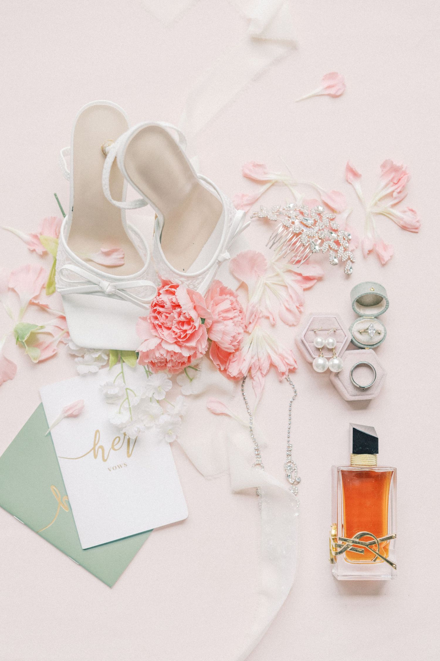 Elegant bridal flat lay with white heels, pink peonies, YSL perfume, pearl jewelry, and ring boxes on blush background.