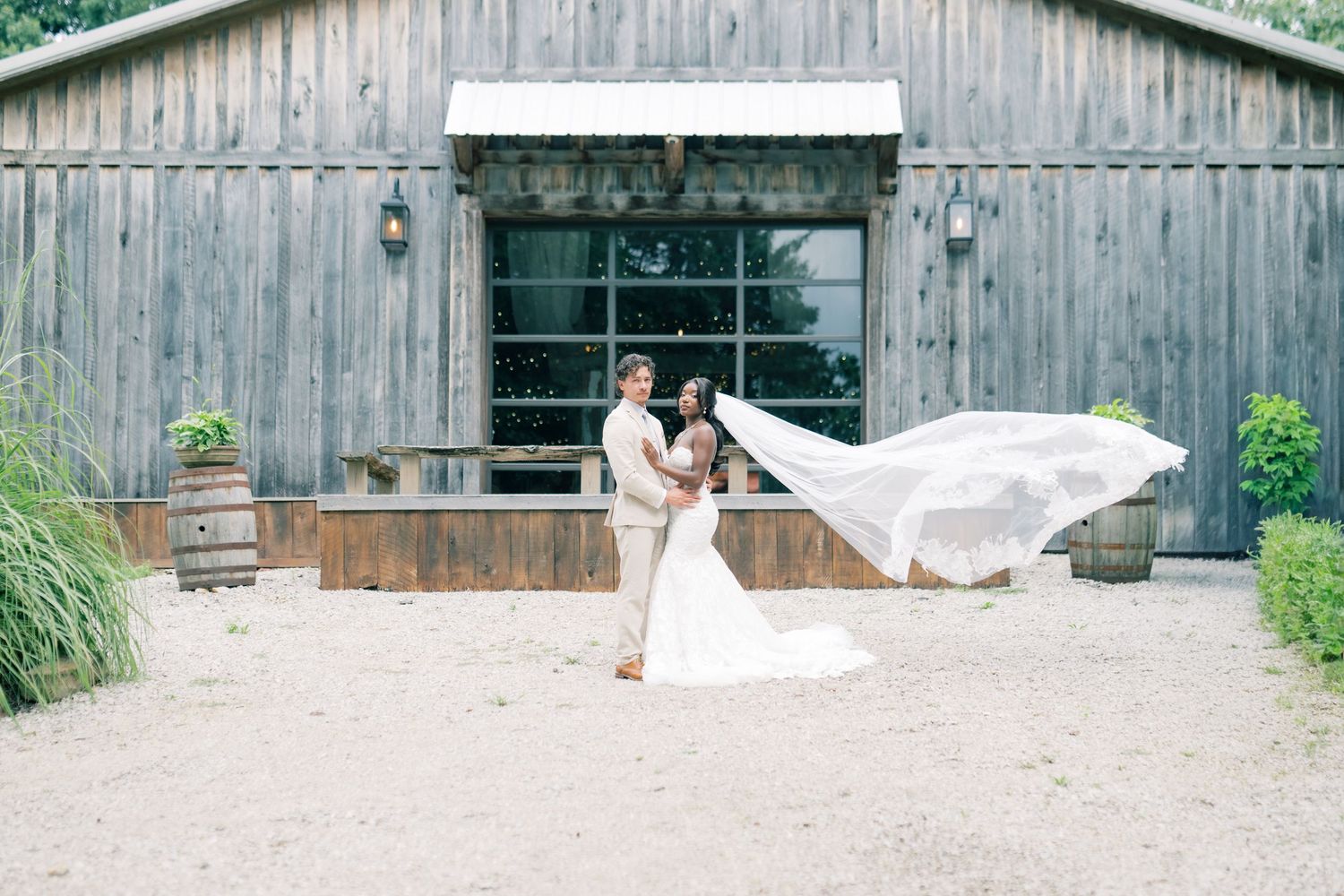 Bride and groom embrace outdoors in front of rustic barn venue with flowing wedding veil billowing in the wind.