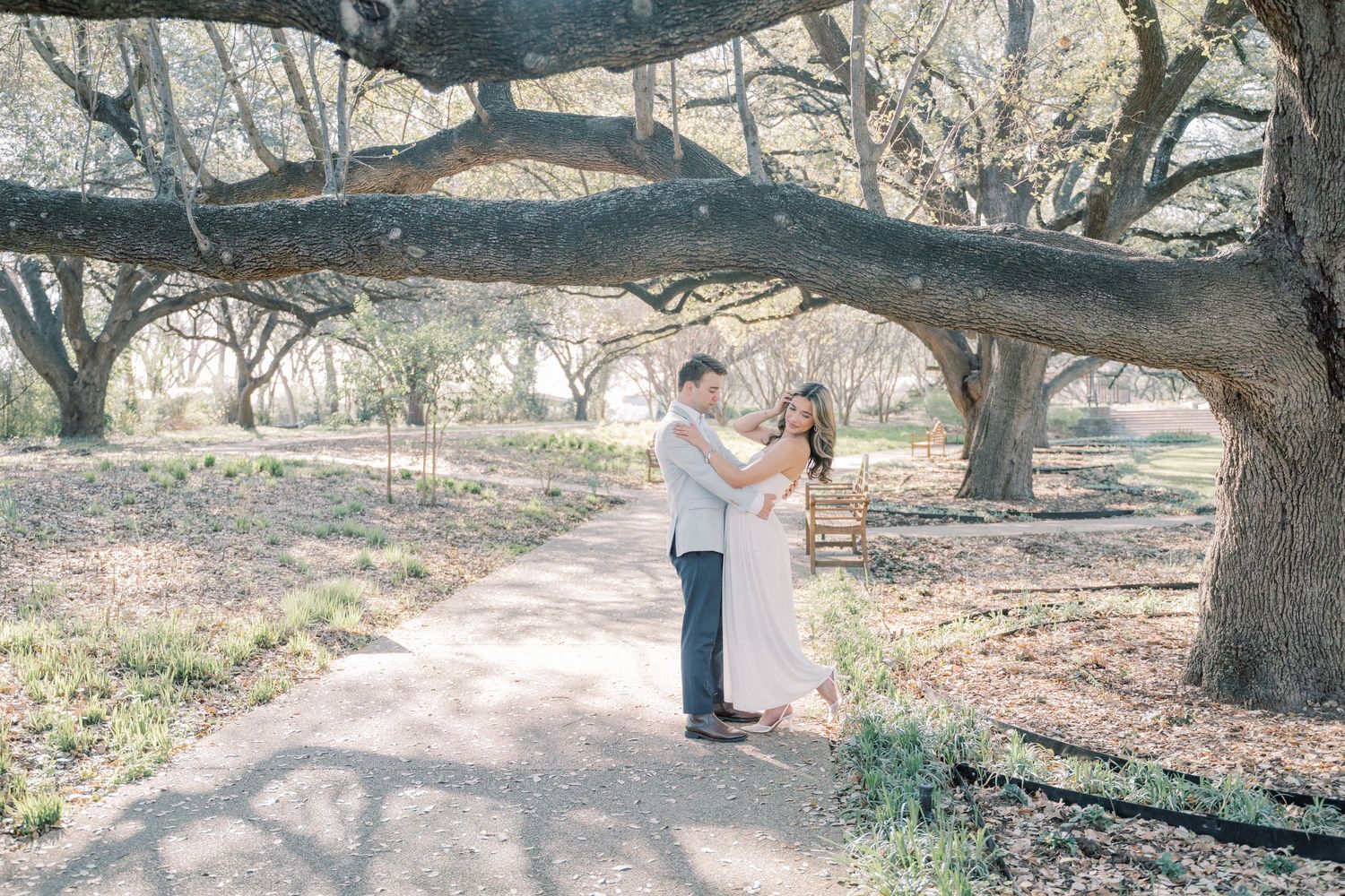 A couple embraces under the branches of a large oak tree in a rustic outdoor setting.