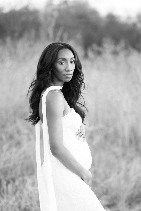 Black and white maternity portrait of an expectant mother in a flowing white dress standing in a field.