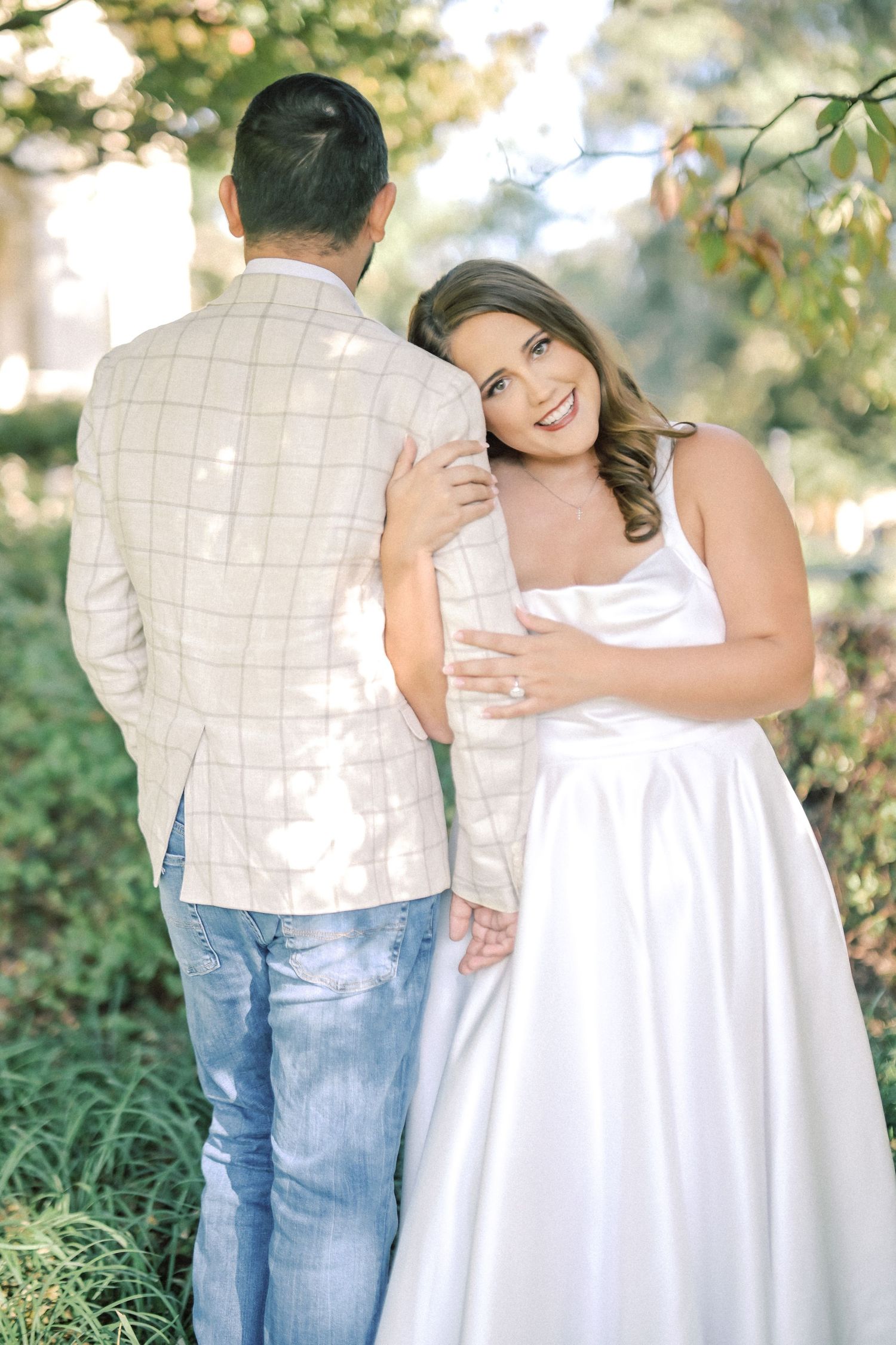 Smiling bride-to-be in a white satin dress leans her head on her fiancé's shoulder as he stands facing away in a plaid blazer.