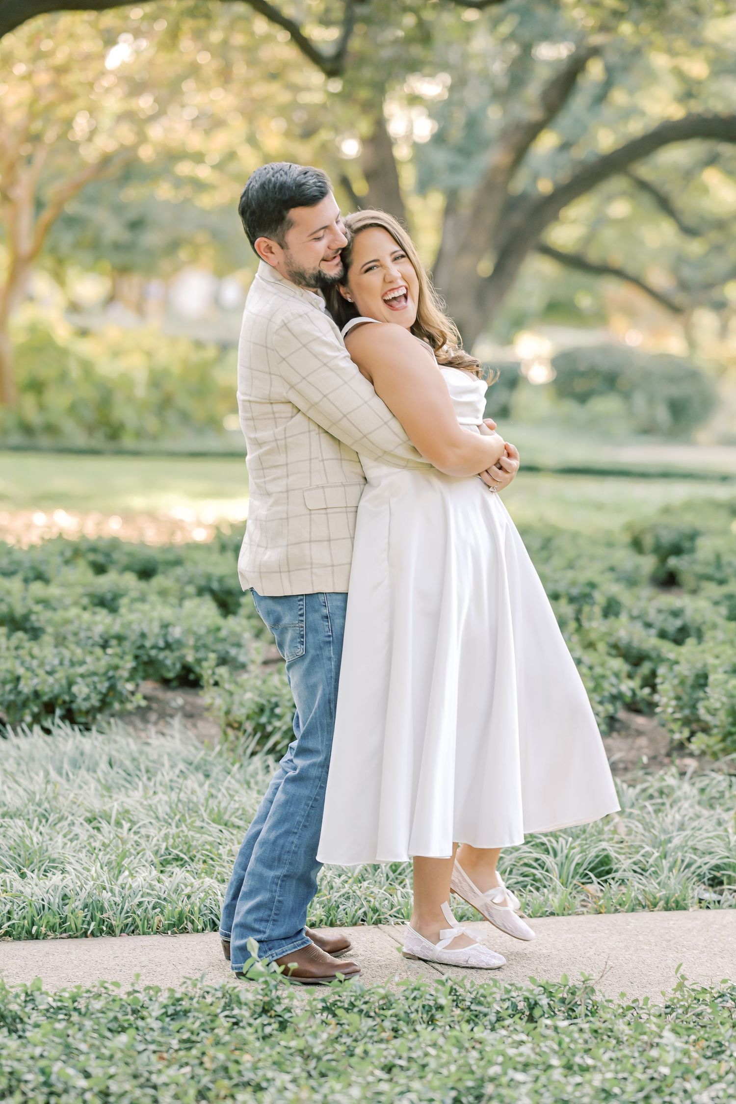 Laughing woman in a white dress is hugged from behind by a smiling man in a plaid blazer on a garden path.