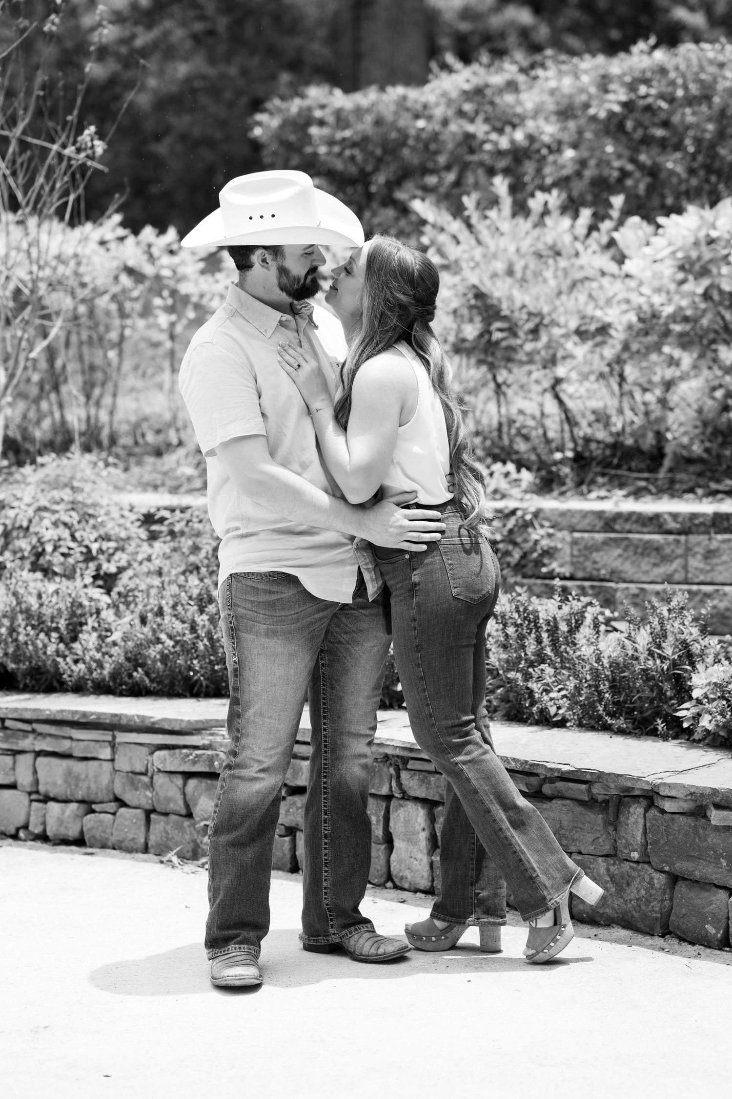 Black and white photo of a couple kissing outdoors, man wearing cowboy hat and jeans, woman in white top.