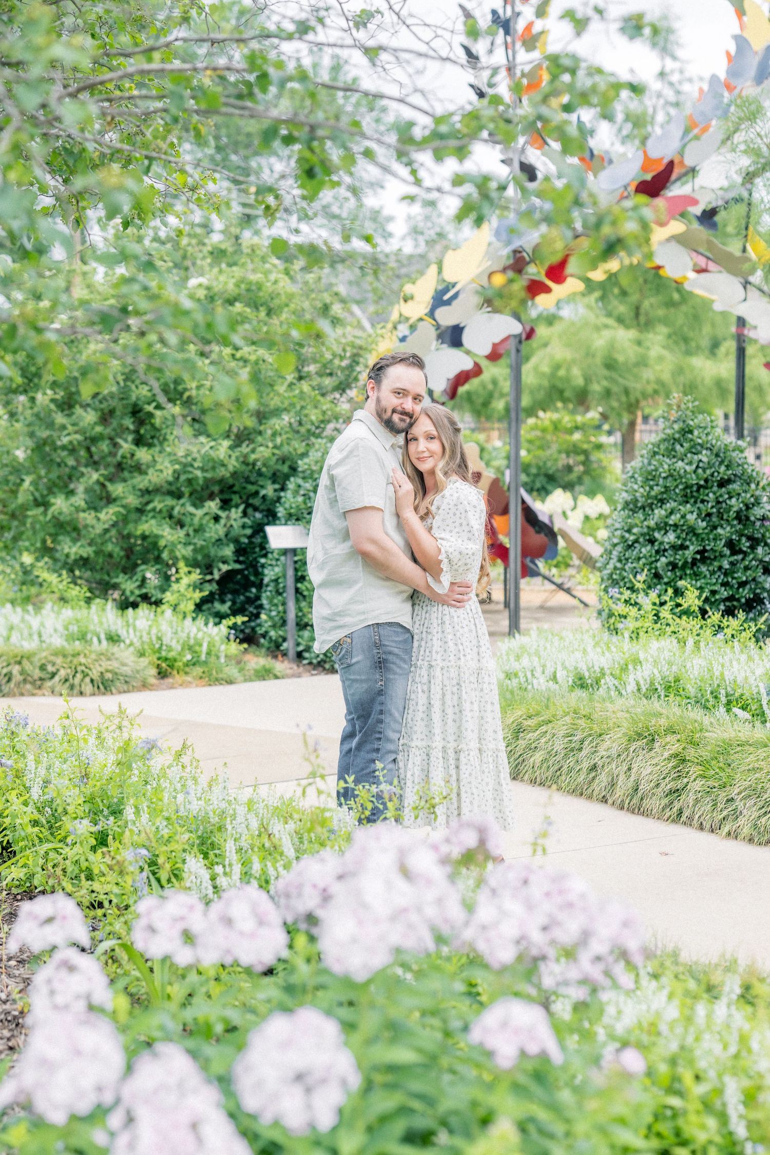 Smiling couple embracing on a garden path with white flowers and butterfly sculpture.