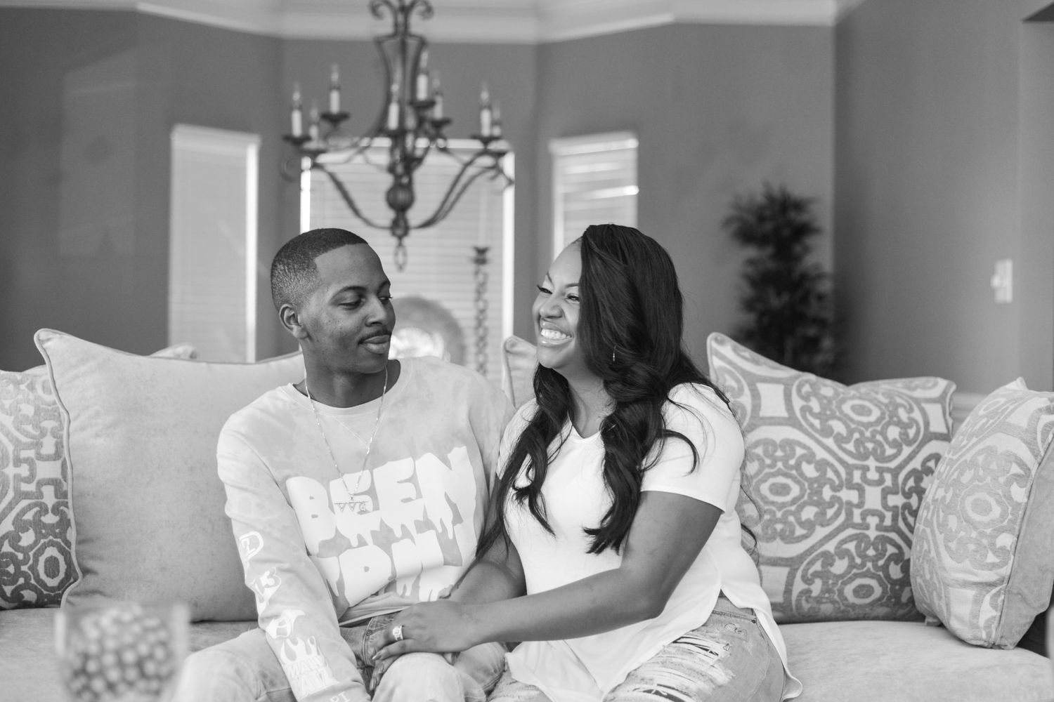 Black and white photo of a couple sitting together on a couch, smiling at each other in a cozy living room.