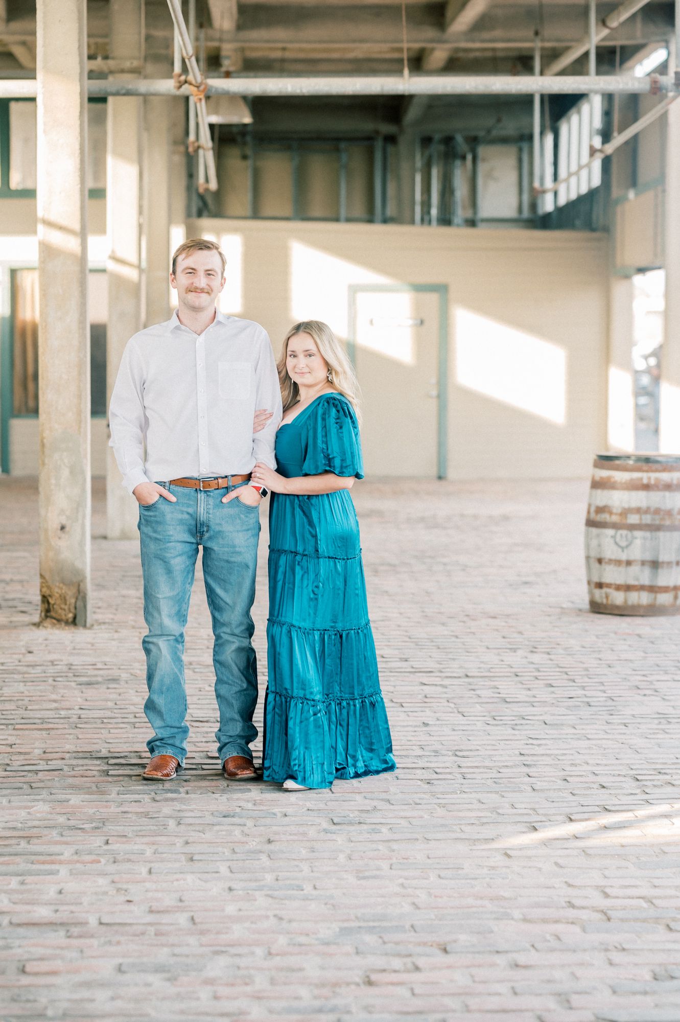 A couple poses together in an industrial space with brick floors and wooden barrels, she wearing a teal tiered dress.