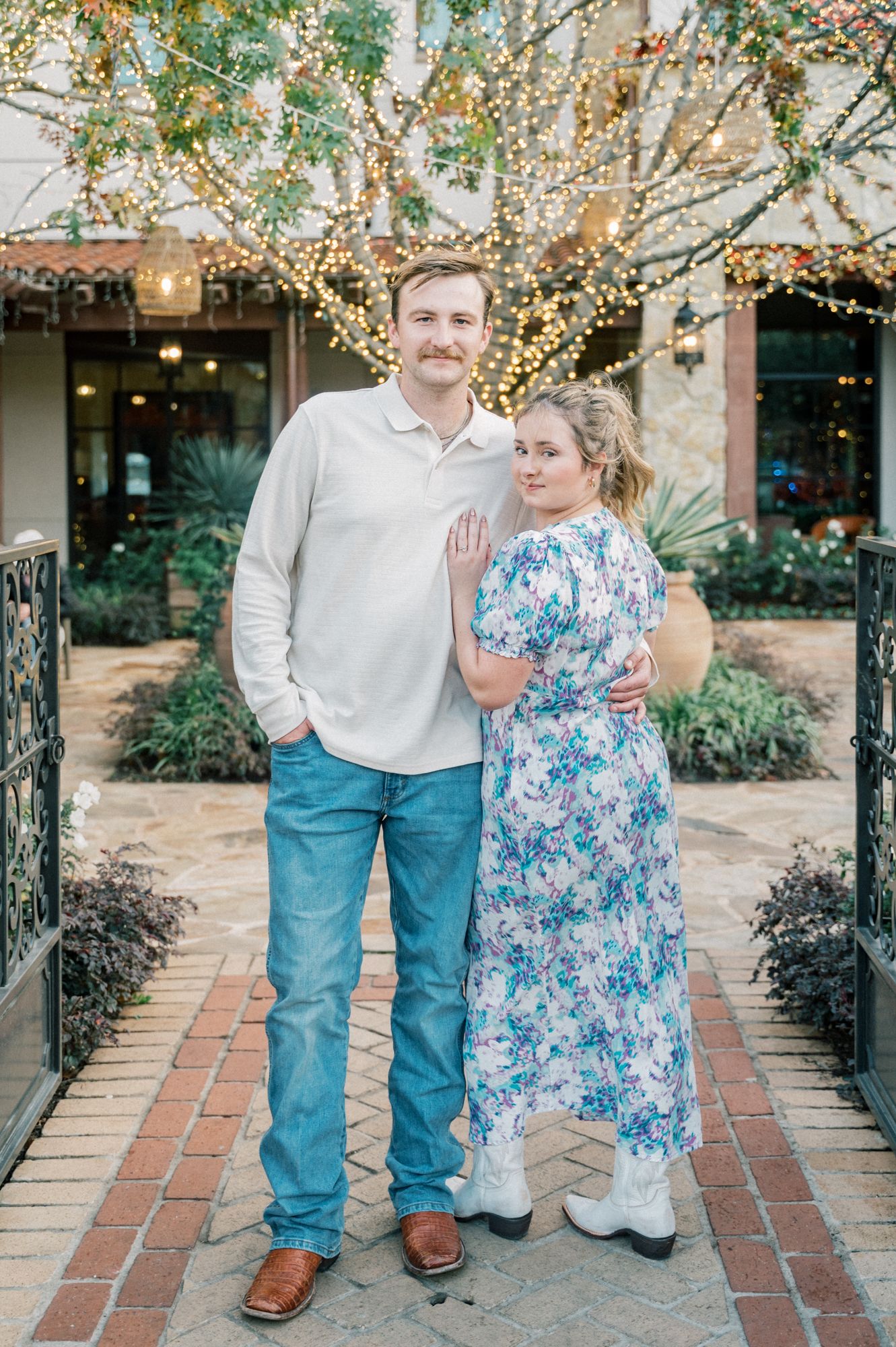A couple poses together outdoors on a brick pathway with decorative lights and trees in the background.