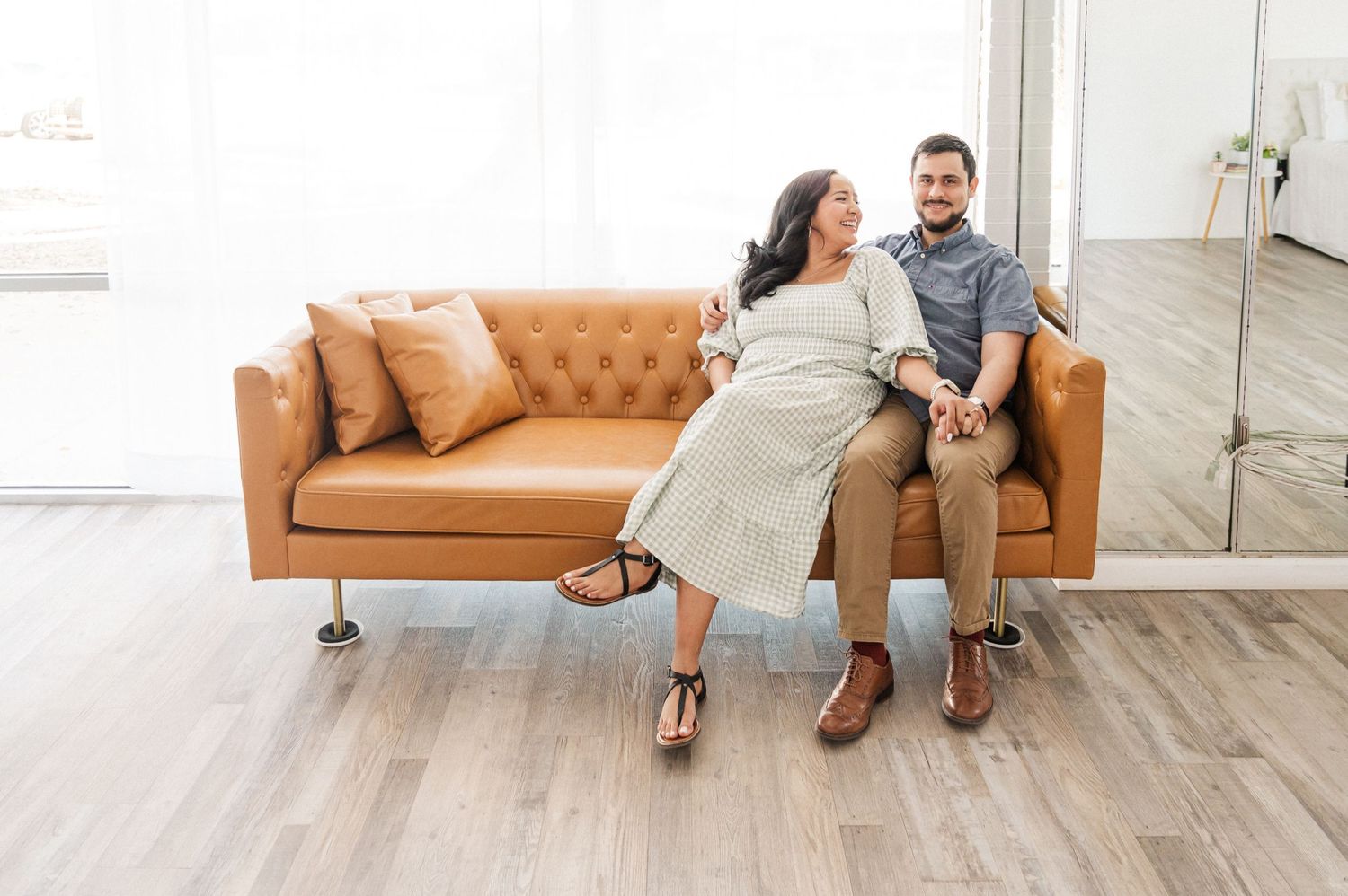 A couple sits together laughing on a tan leather tufted sofa in a bright modern room with wood floors.