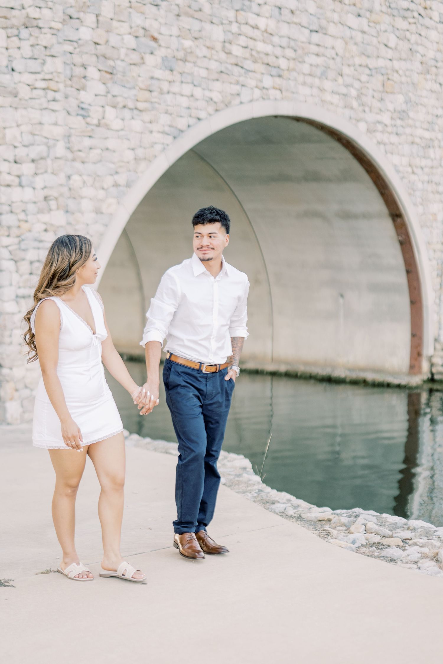 Couple holding hands smiling at each other near stone arch bridge and water.