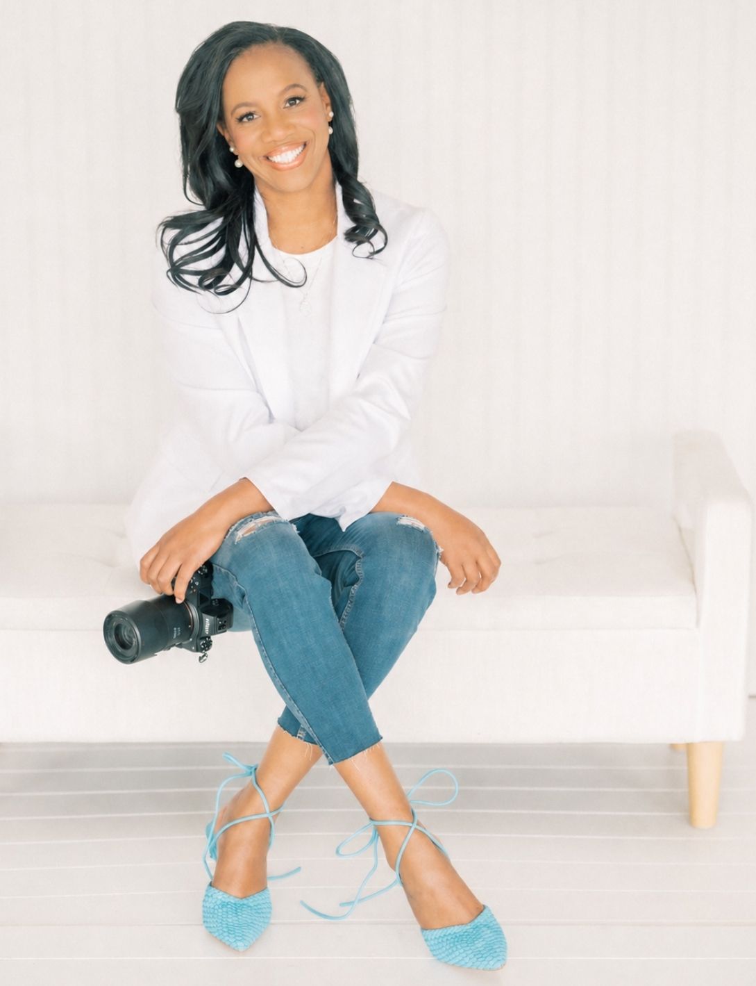 Smiling woman in white blazer and jeans sits on white couch holding a camera, wearing teal heels.