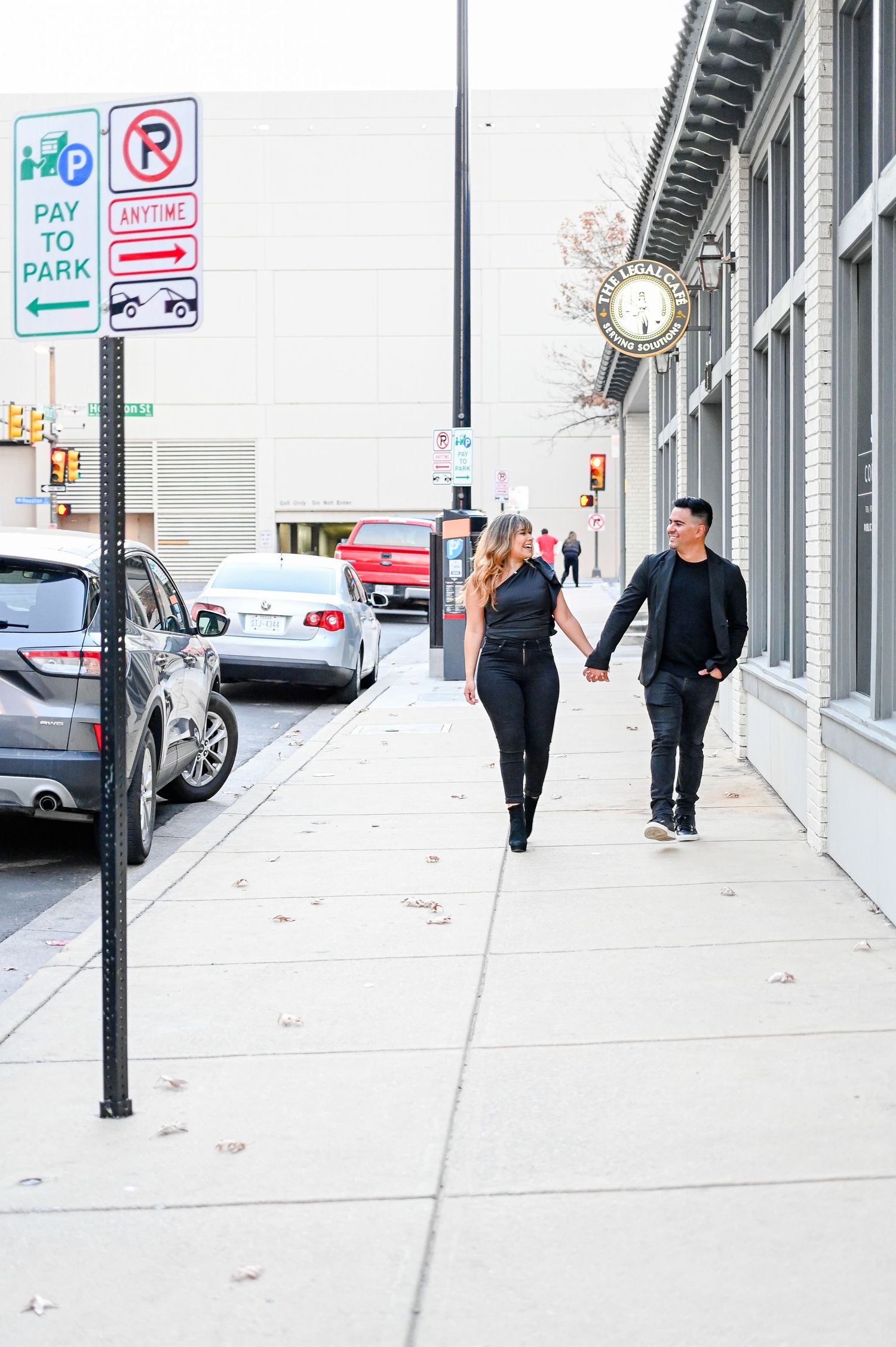 Stylish couple holding hands and strolling along a downtown sidewalk with parking signs nearby.