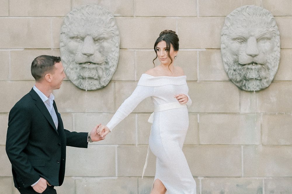 Pregnant woman in white off-shoulder dress holds hands with man in dark suit near stone lion wall sculptures.
