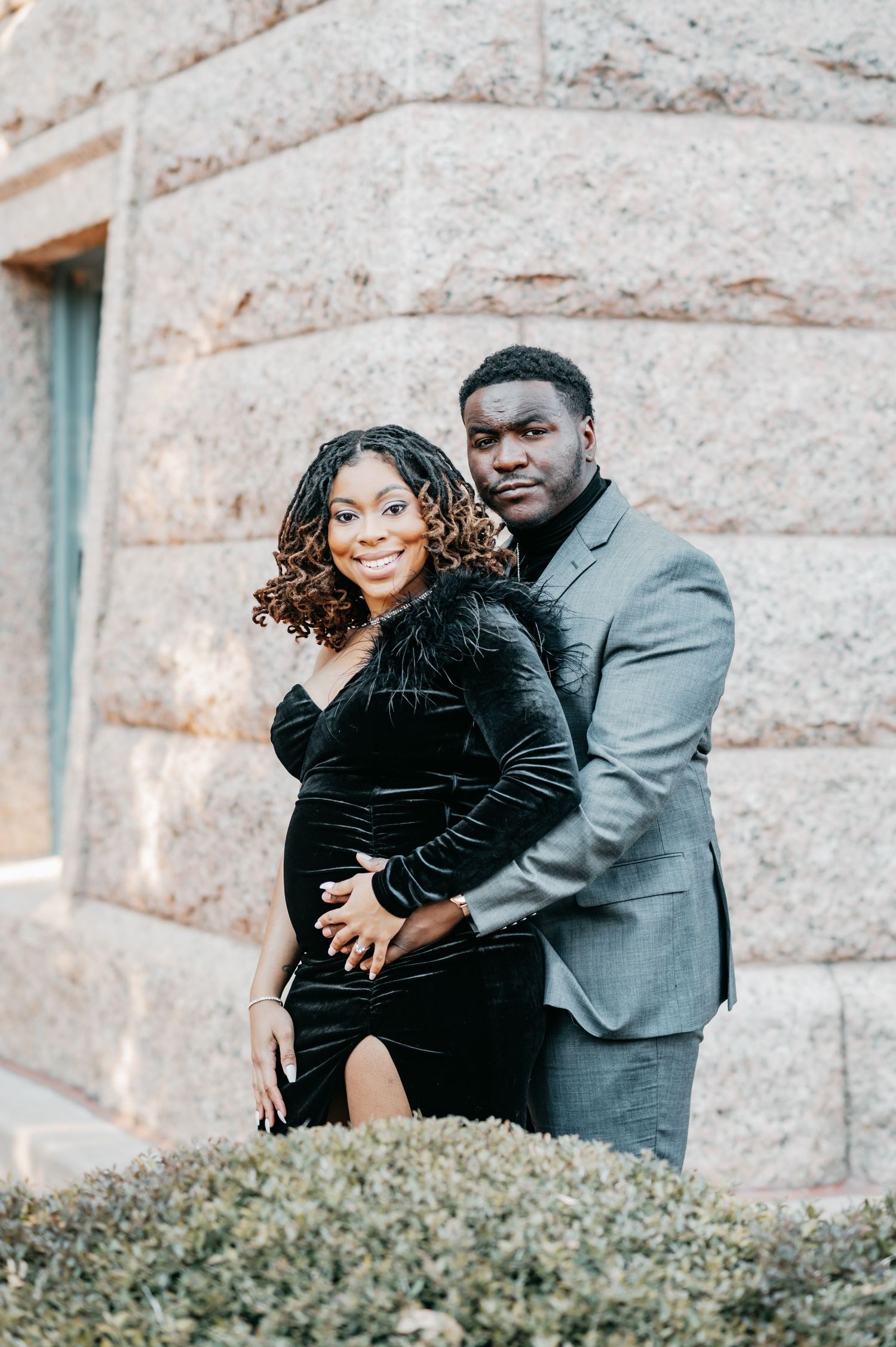 Couple posing for maternity photos outdoors, woman in black velvet dress, man in gray suit, against stone wall.
