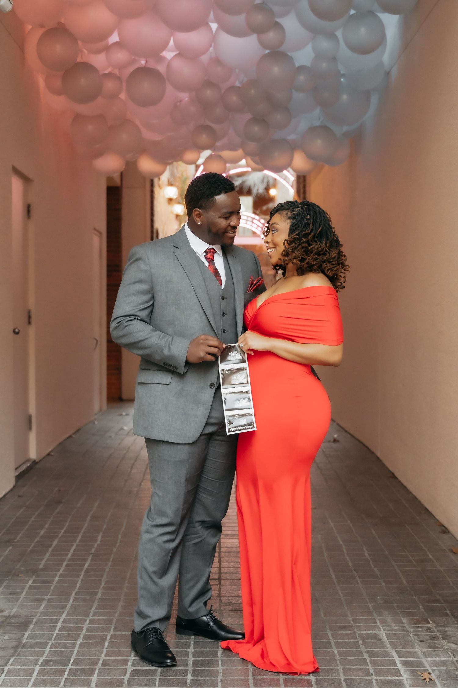 Couple in elegant attire pose in balloon-filled hallway, woman in red maternity gown holding ultrasound photo.