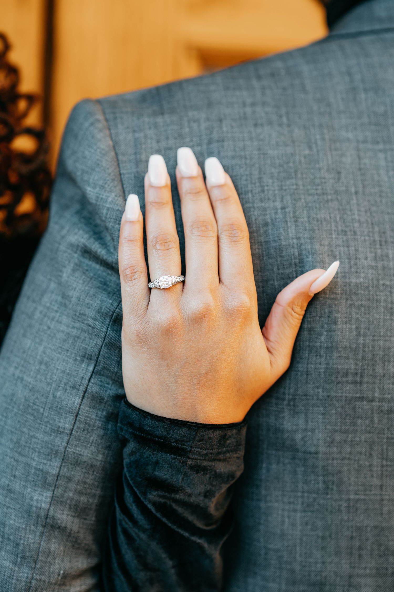 Close-up of a diamond engagement ring on a womans hand resting on a gray suit jacket.