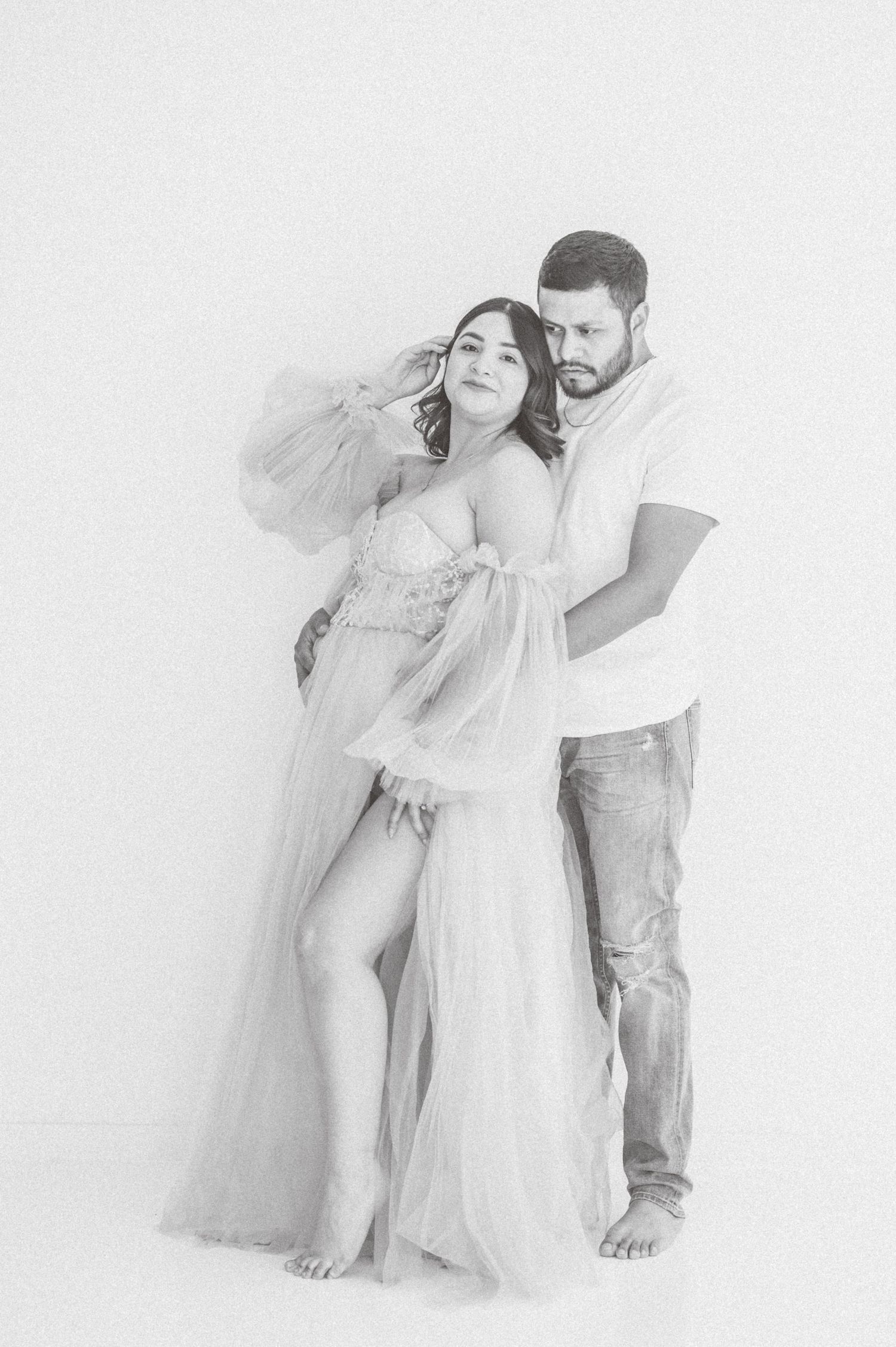 Black and white maternity photo of a couple embracing, woman in flowing tulle gown, man in casual jeans and t-shirt.