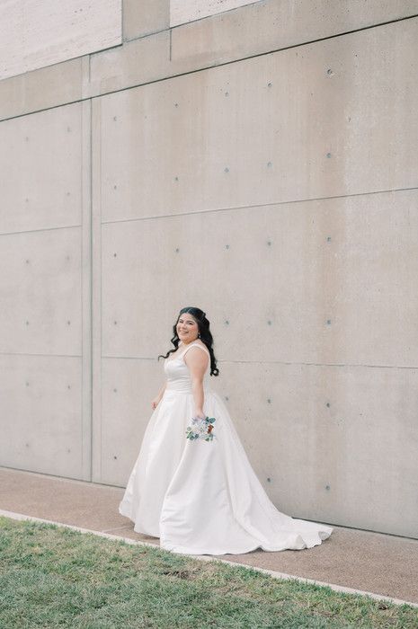 Bride in a white ball gown smiling and walking along a concrete wall while holding a bouquet.