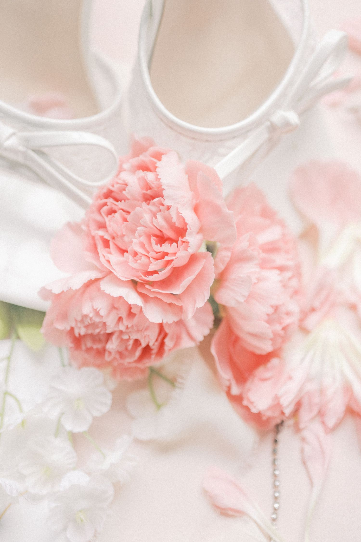 Pink carnations rest beside white bridal heels on a soft pastel background, perfect for wedding decor inspiration.
