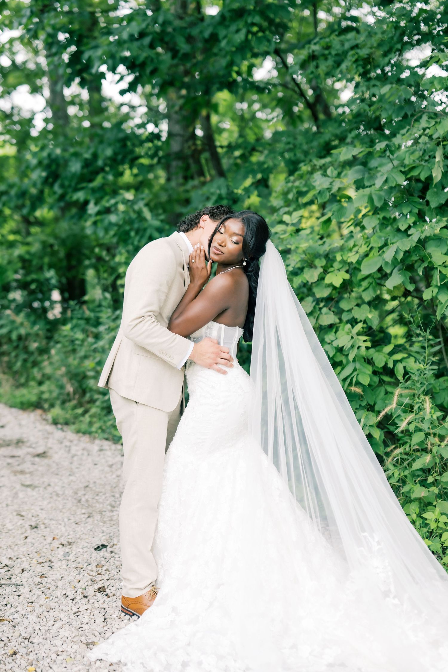 Groom in tan suit tenderly kisses bride in lace mermaid gown among lush green trees.