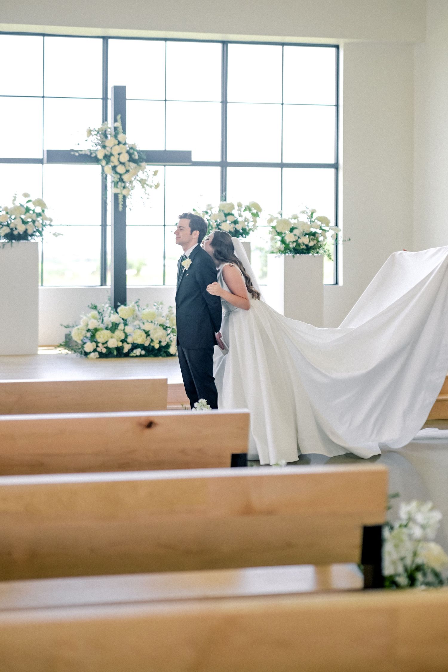 Bride and groom embrace at church altar with white floral arrangements and cross during wedding ceremony.