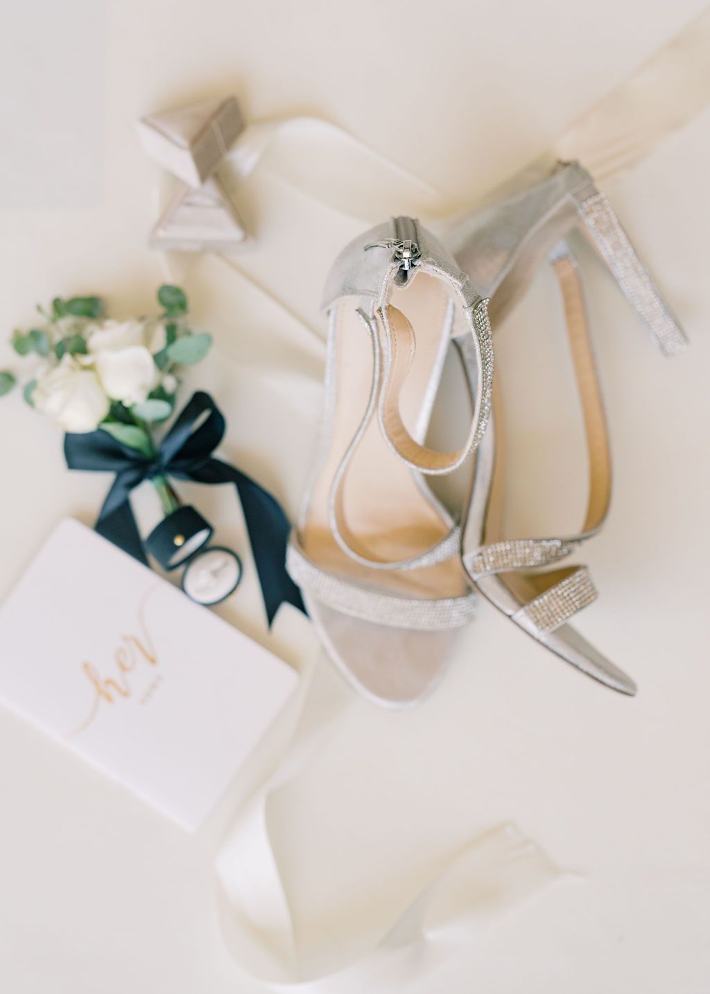 Silver glitter bridal heels with ankle strap beside white roses, navy ribbon, and monogrammed wedding stationery.