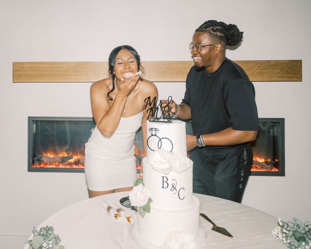 Couple cutting a white wedding cake decorated with B&C monogram and floral accents near a modern fireplace.