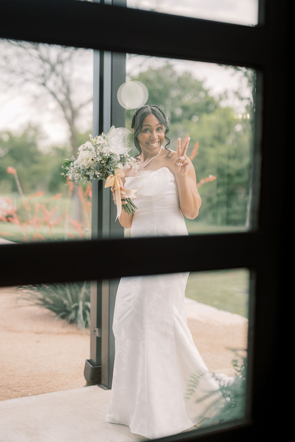 Smiling bride in white gown holds bouquet and flashes peace sign, photographed through glass door at outdoor venue.