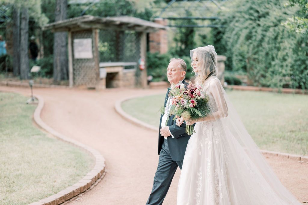 Bride in lace gown with veil holds bouquet beside suited man on garden path with greenery and brick pavilion.