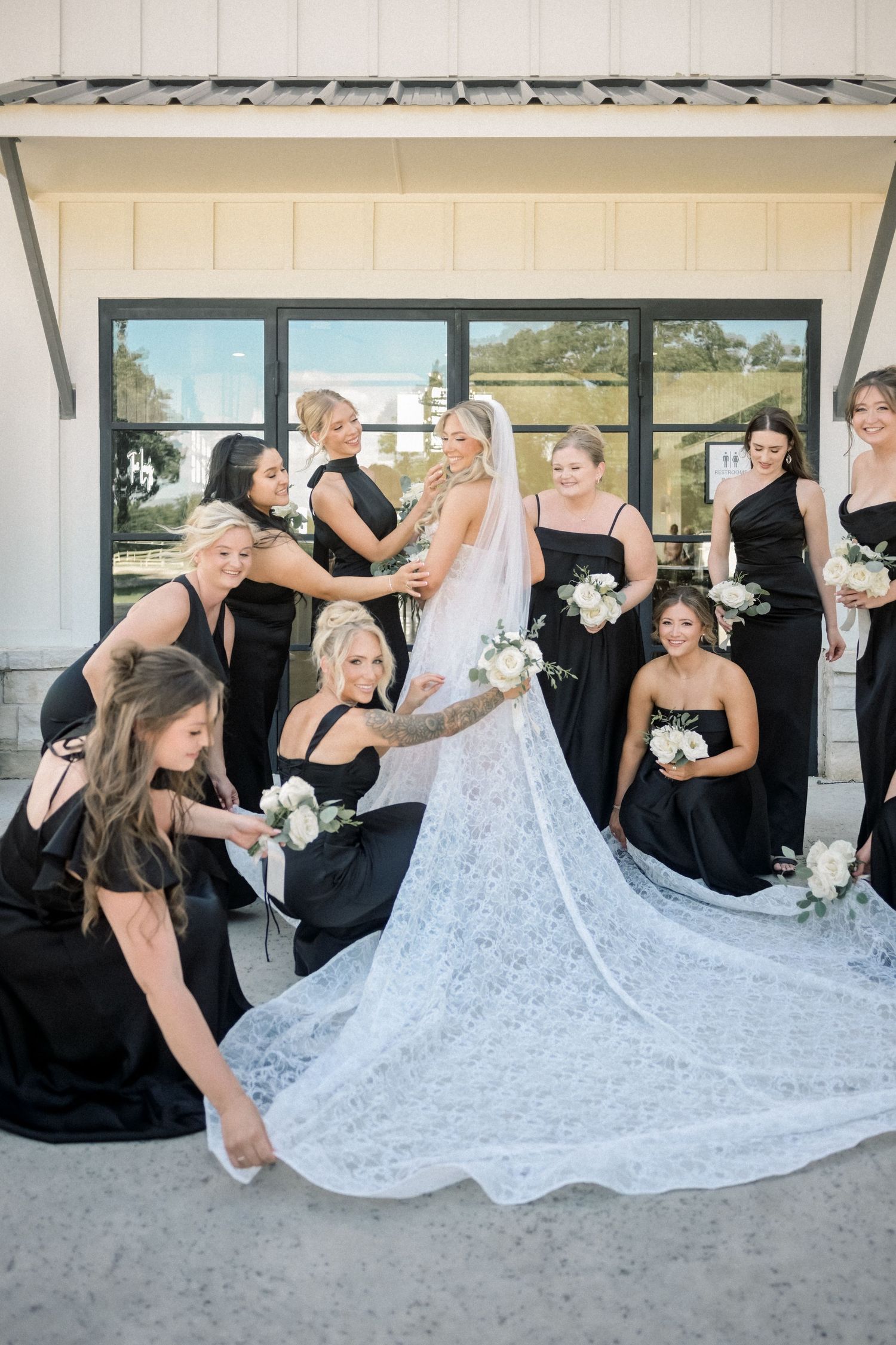 Bride in lace gown surrounded by bridesmaids in black dresses arranging her veil and train outside.