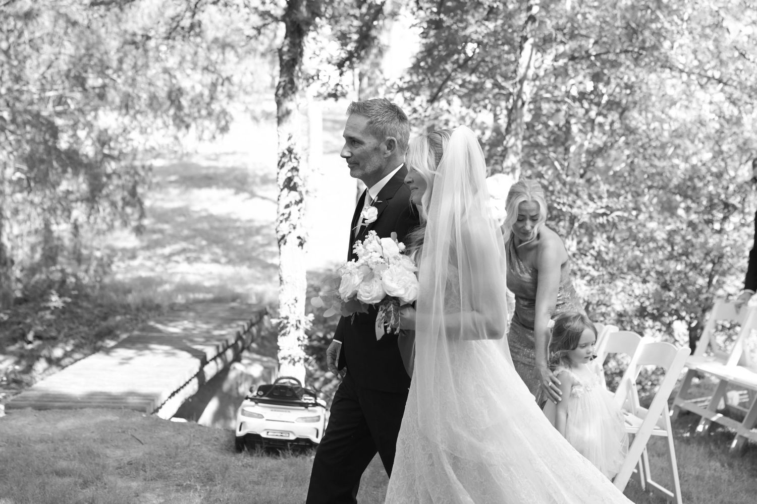 Black and white photo of bride walking down aisle with father during outdoor wedding ceremony.
