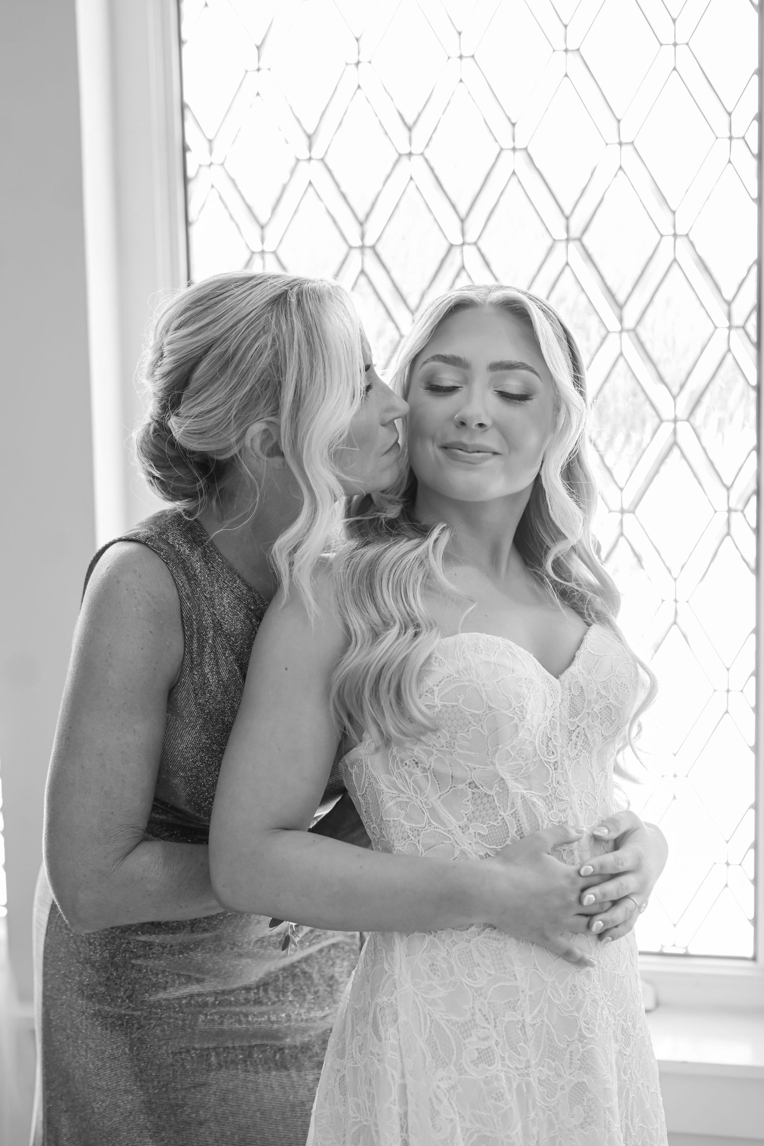 Black and white photo of mother kissing bride on cheek while embracing her in wedding dress.