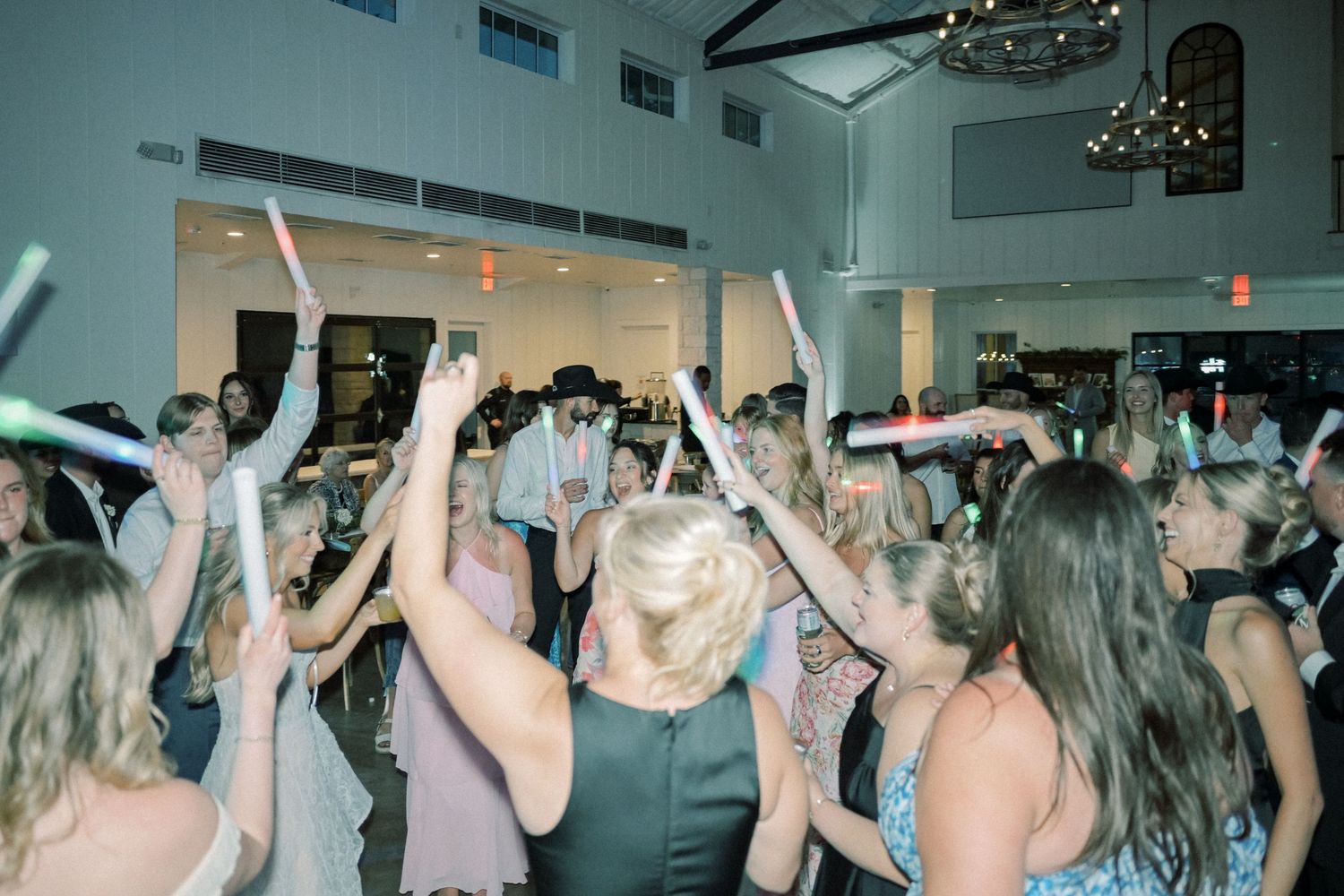 Energetic wedding reception dance floor with guests raising glow sticks in bright white venue.