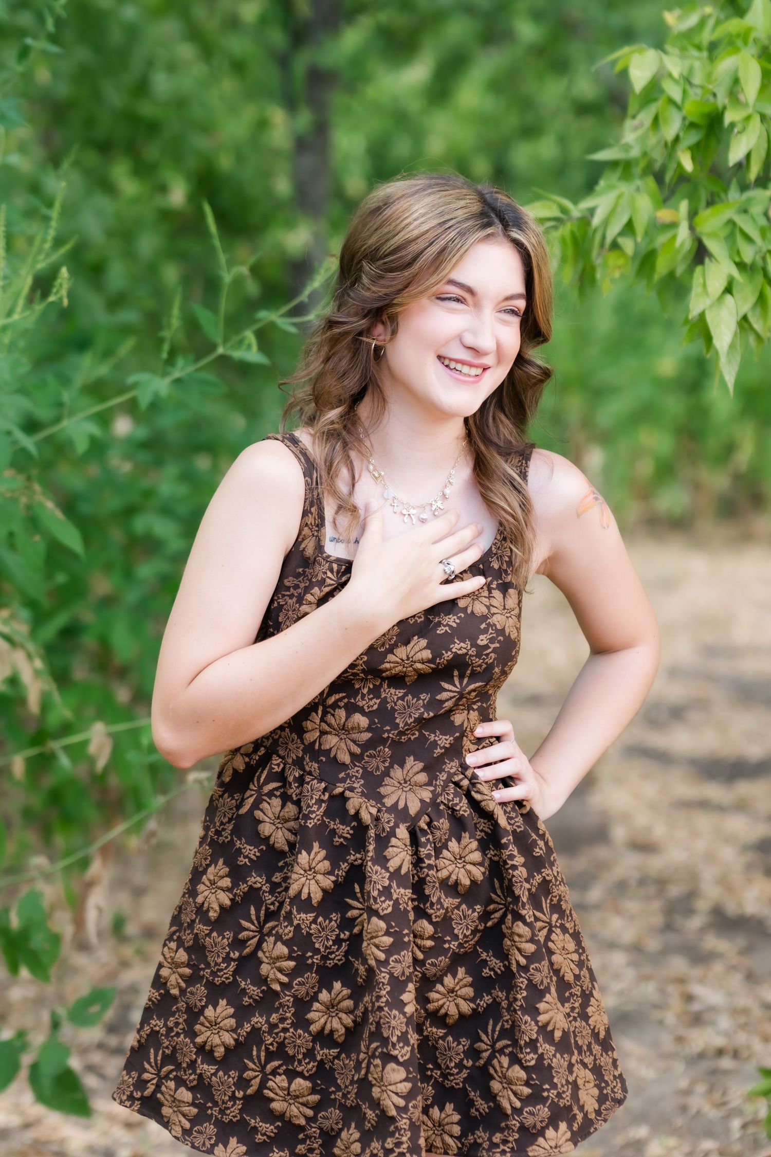 Young woman in brown floral dress smiling outdoors among green trees, hand on chest, wearing silver necklace.