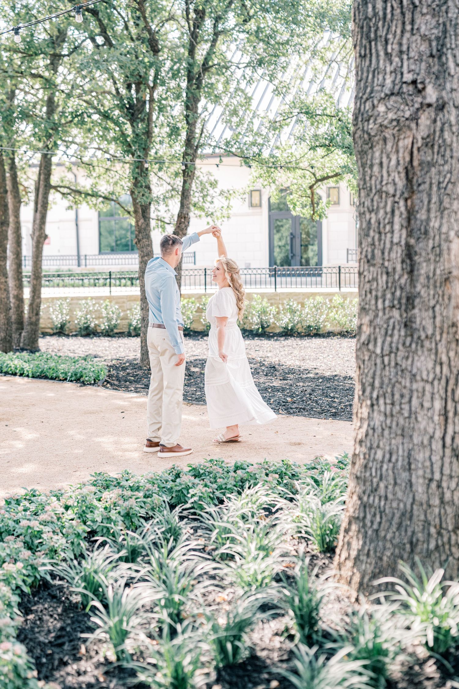 Couple dancing together in a lush garden, man spinning woman in flowing white dress under tall trees.