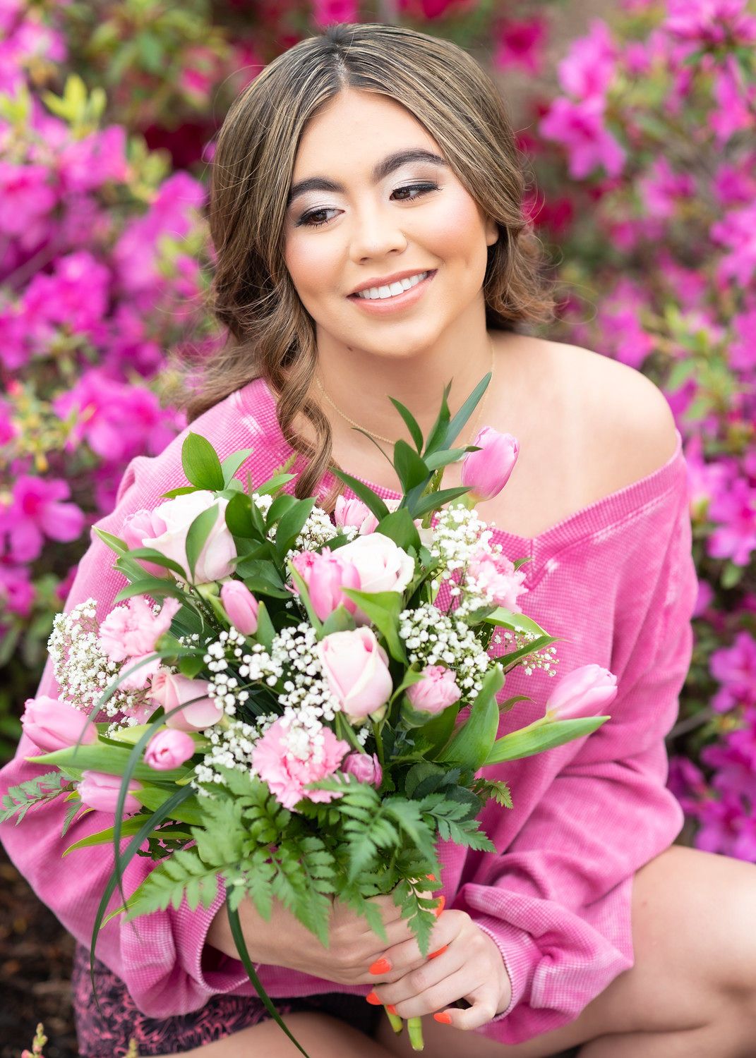Young woman in pink off-shoulder sweater holding bouquet of pink tulips and roses, smiling among blooming azaleas.