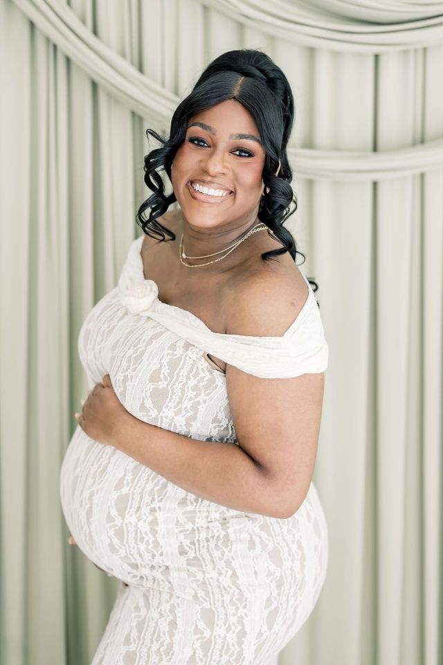 Smiling pregnant woman in white lace off-shoulder maternity gown embracing baby bump during elegant photo shoot.