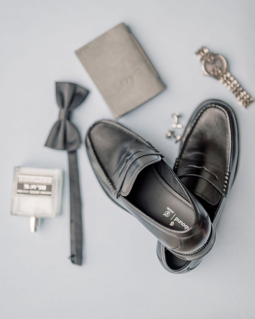 Groom wedding accessories flat lay featuring black loafers, bow tie, watch, cufflinks, and wallet.