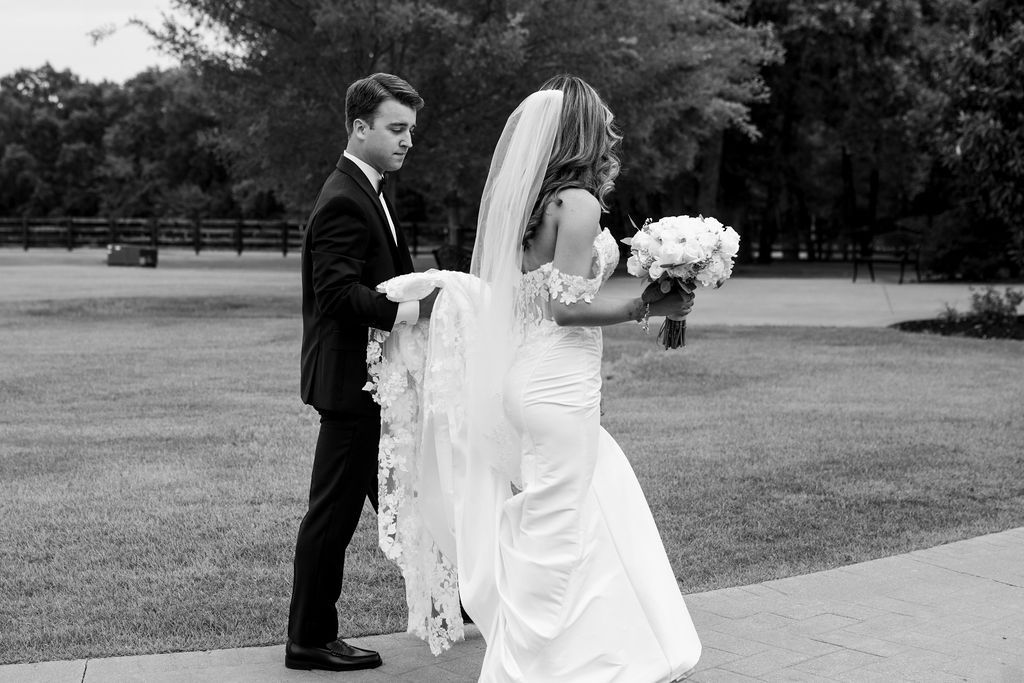Bride and groom walk together outdoors, black and white wedding photo with veil trailing behind.