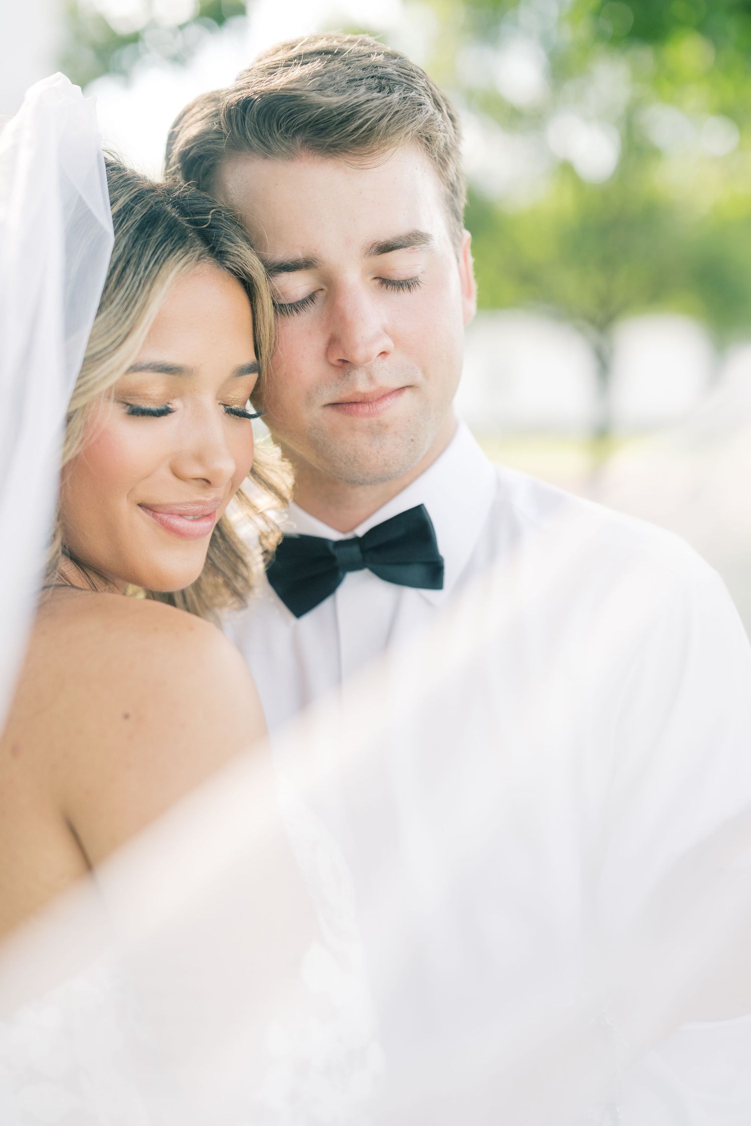 Bride and groom share a tender moment, eyes closed, as her veil drapes softly around them outdoors.