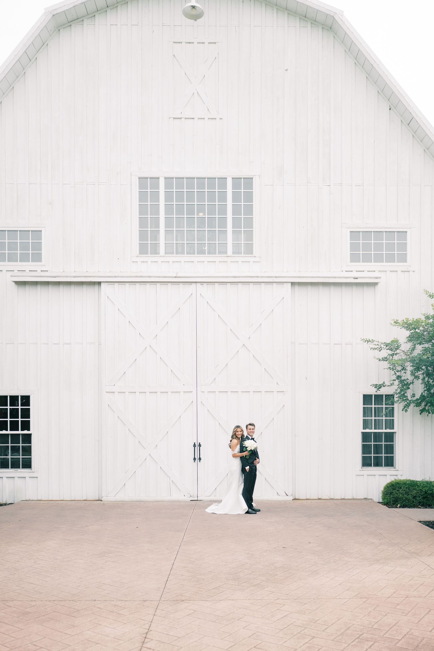 Bride and groom posing in front of a large white barn with double doors on their wedding day.