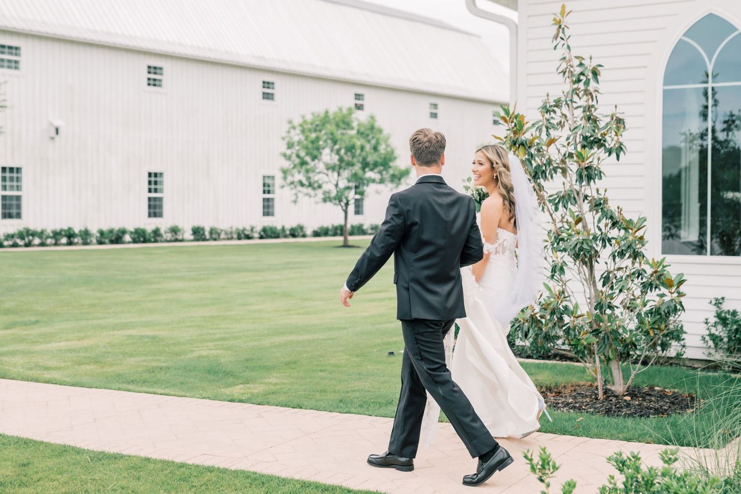 Bride and groom walking hand in hand across a green lawn outside a white wedding venue.