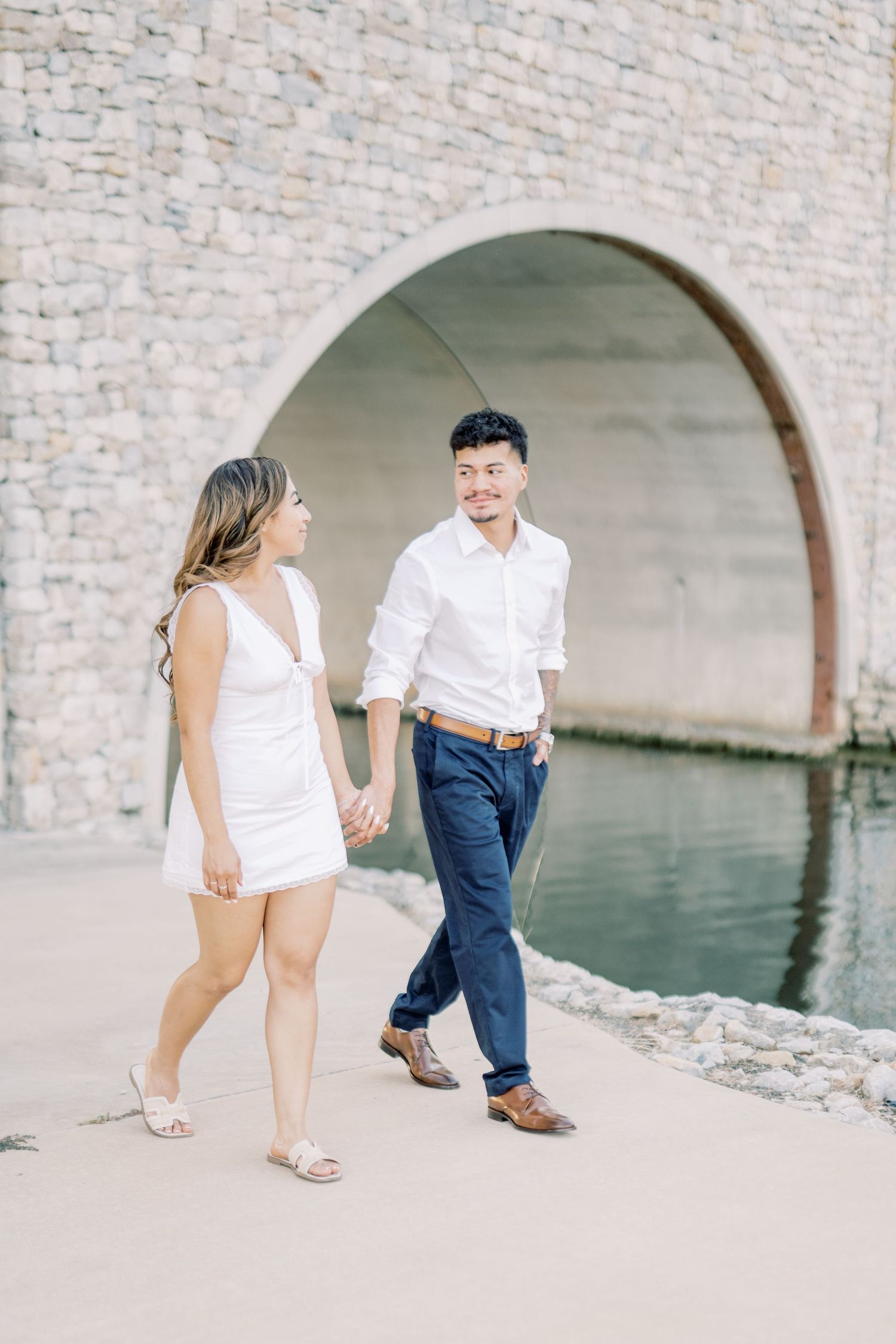 A smiling couple in white outfits holds hands while strolling near a stone arch and reflecting water.